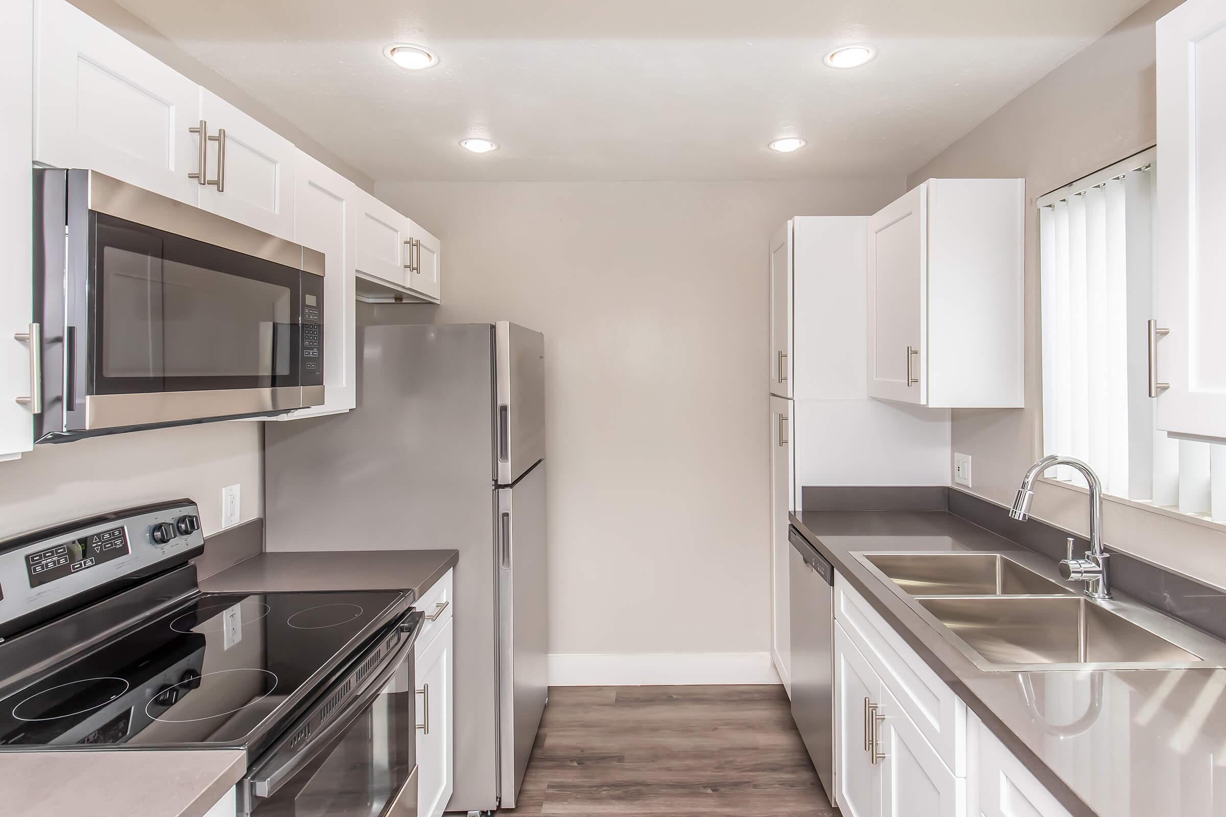 Modern kitchen featuring white cabinets, stainless steel appliances including a microwave, refrigerator, and stove. The countertops are dark and the space is well-lit with recessed lighting. A double sink is positioned near a window with vertical blinds, showcasing a clean and contemporary design.