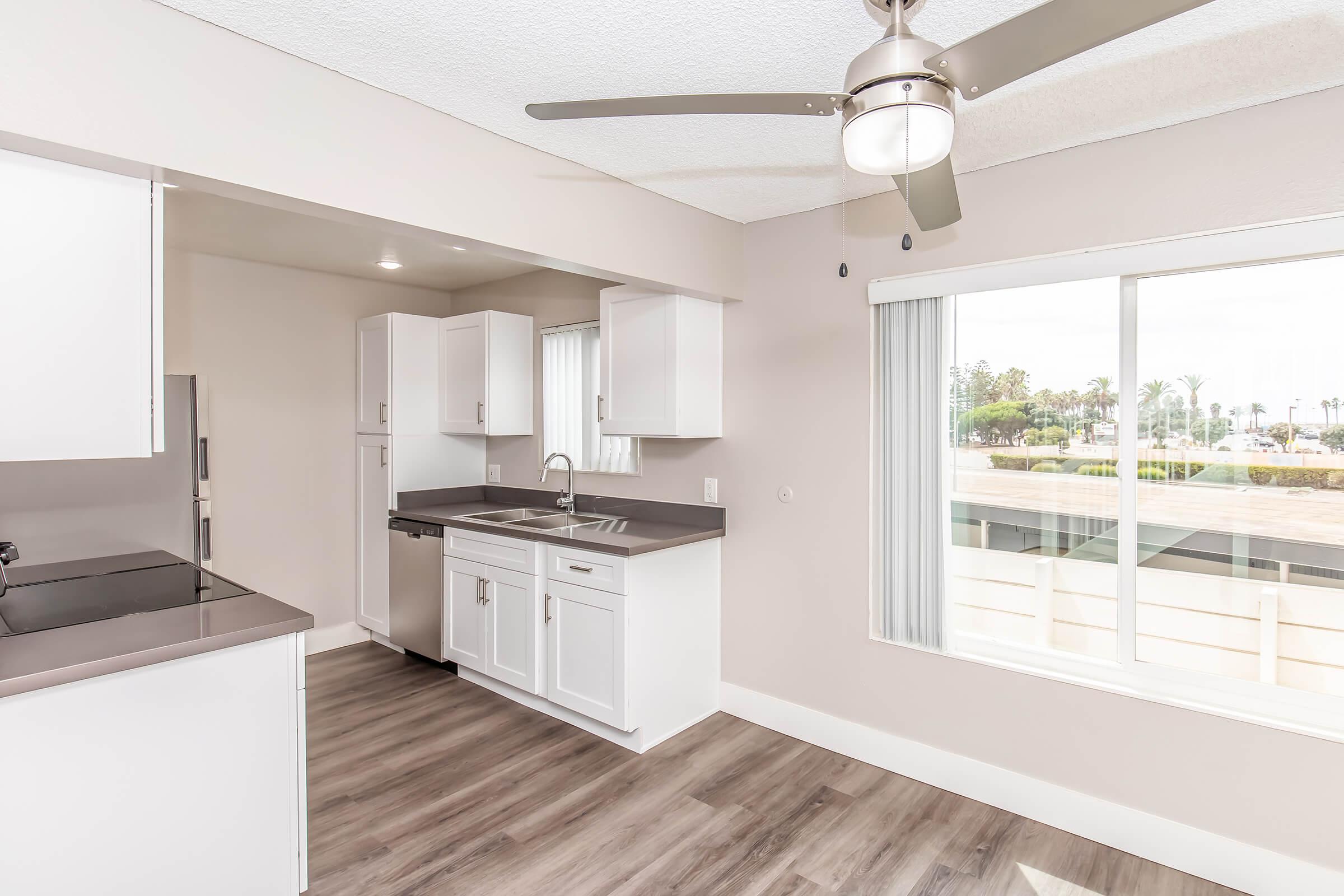 A bright, modern kitchen with white cabinets and black countertops. The space features a ceiling fan, large window with vertical blinds, and a view of a palm tree-lined exterior. The flooring is light wood, and the kitchen area is well-lit with natural light.