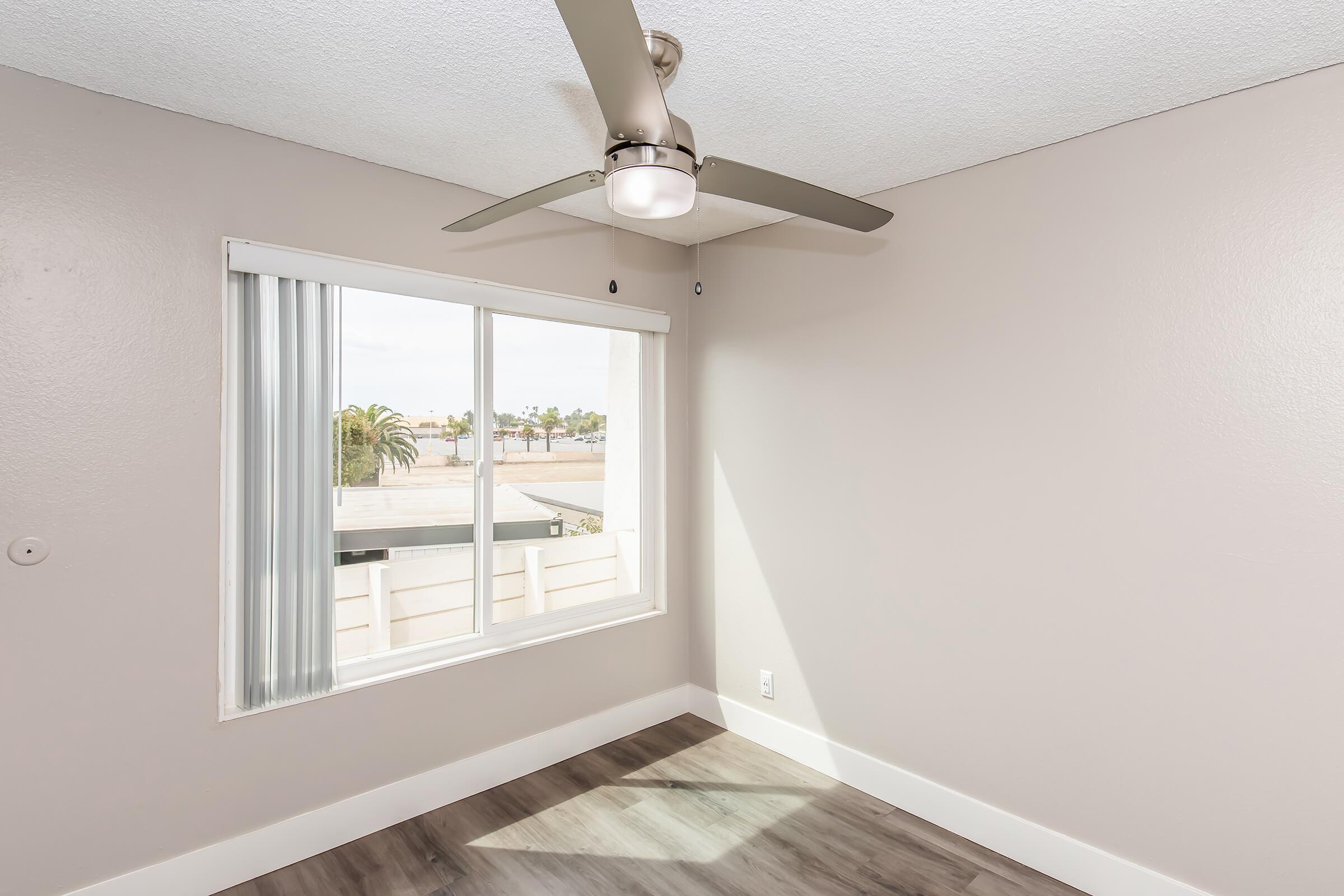 A well-lit empty room featuring a modern ceiling fan, light gray walls, and a window with vertical blinds. The view outside includes palm trees and a clear sky. The floor is a light-colored laminate, enhancing the bright and airy feel of the space.