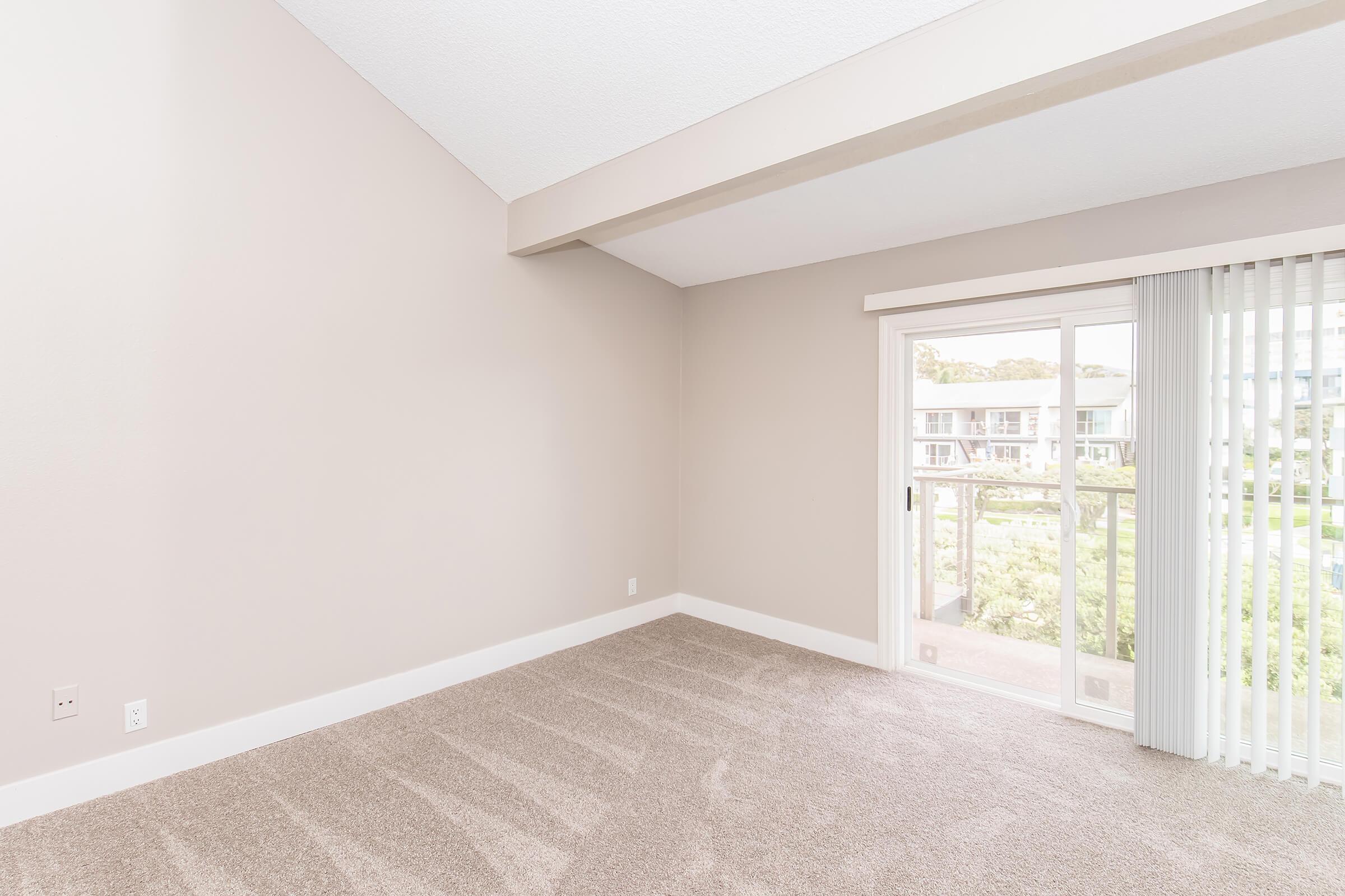 Spacious empty room with light beige walls and carpet, featuring a large sliding glass door that opens to a balcony with a view of greenery outside. Natural light floods in from the door, creating a bright and airy atmosphere.