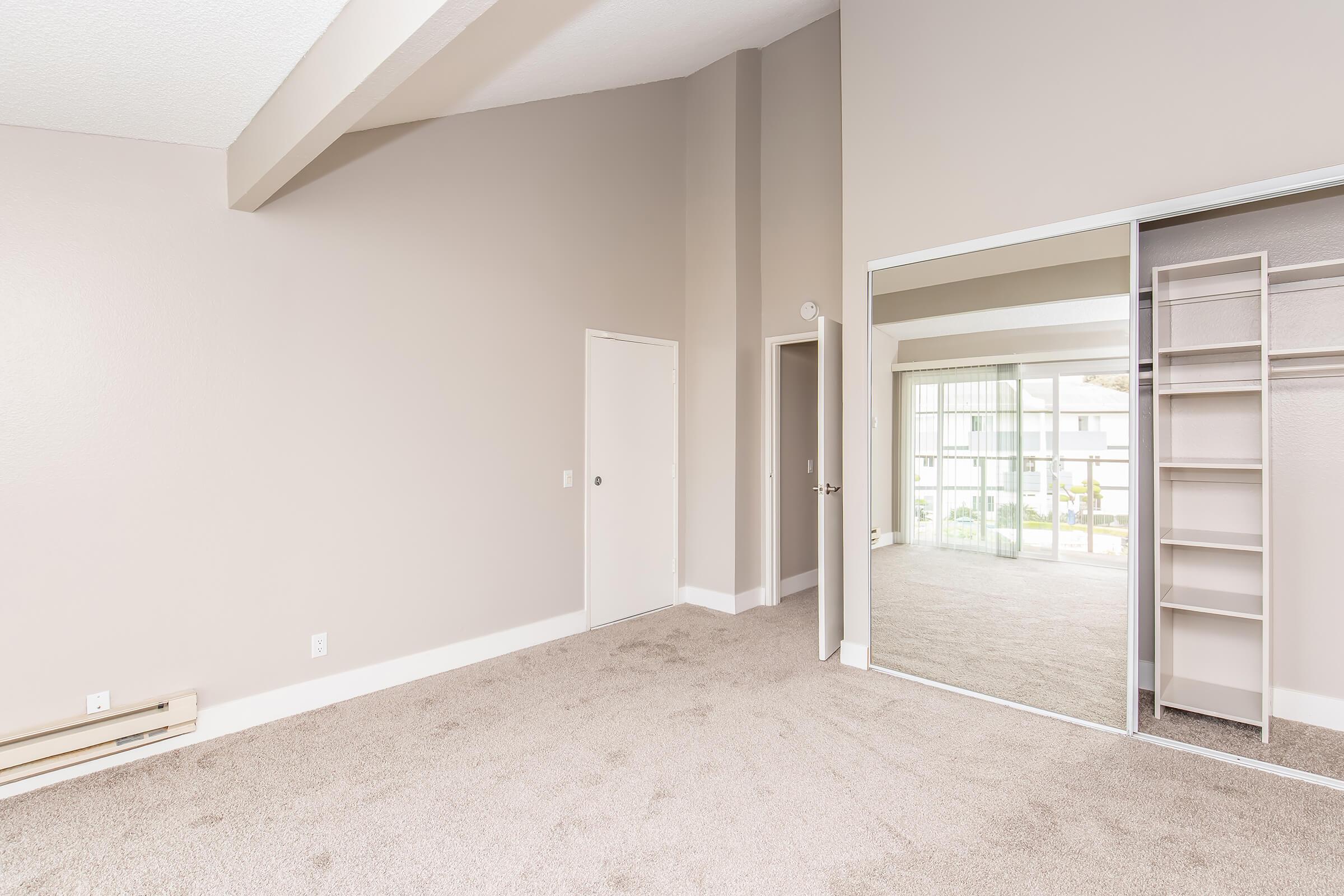 A spacious, well-lit room featuring beige walls and carpet, with high ceilings. It includes a mirrored closet door and another door leading to a hallway. A large window allows natural light to enter, and there are built-in shelves in the closet. The overall atmosphere is bright and inviting.