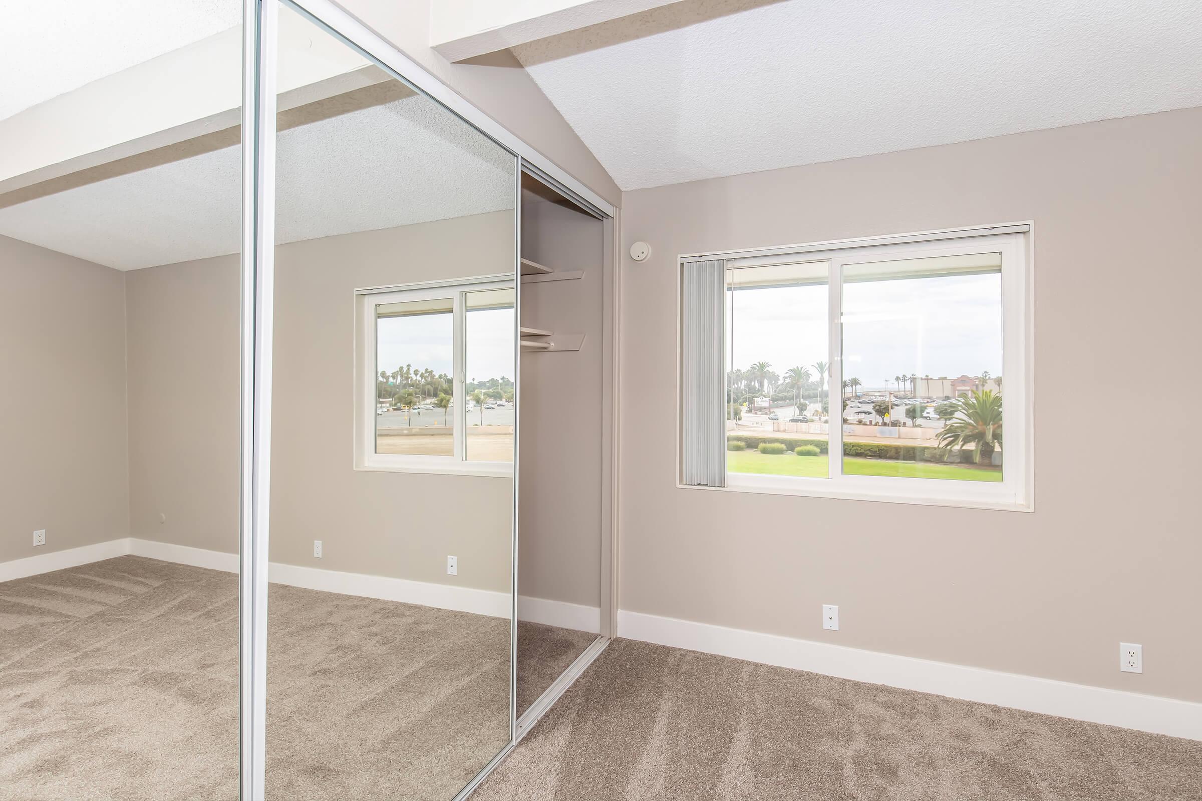 A spacious room featuring a mirrored closet on one wall, with light beige walls and plush carpet. A window on the opposite wall offers views of greenery and buildings outside, allowing natural light to illuminate the space. The ceiling is textured and the overall aesthetic is modern and inviting.