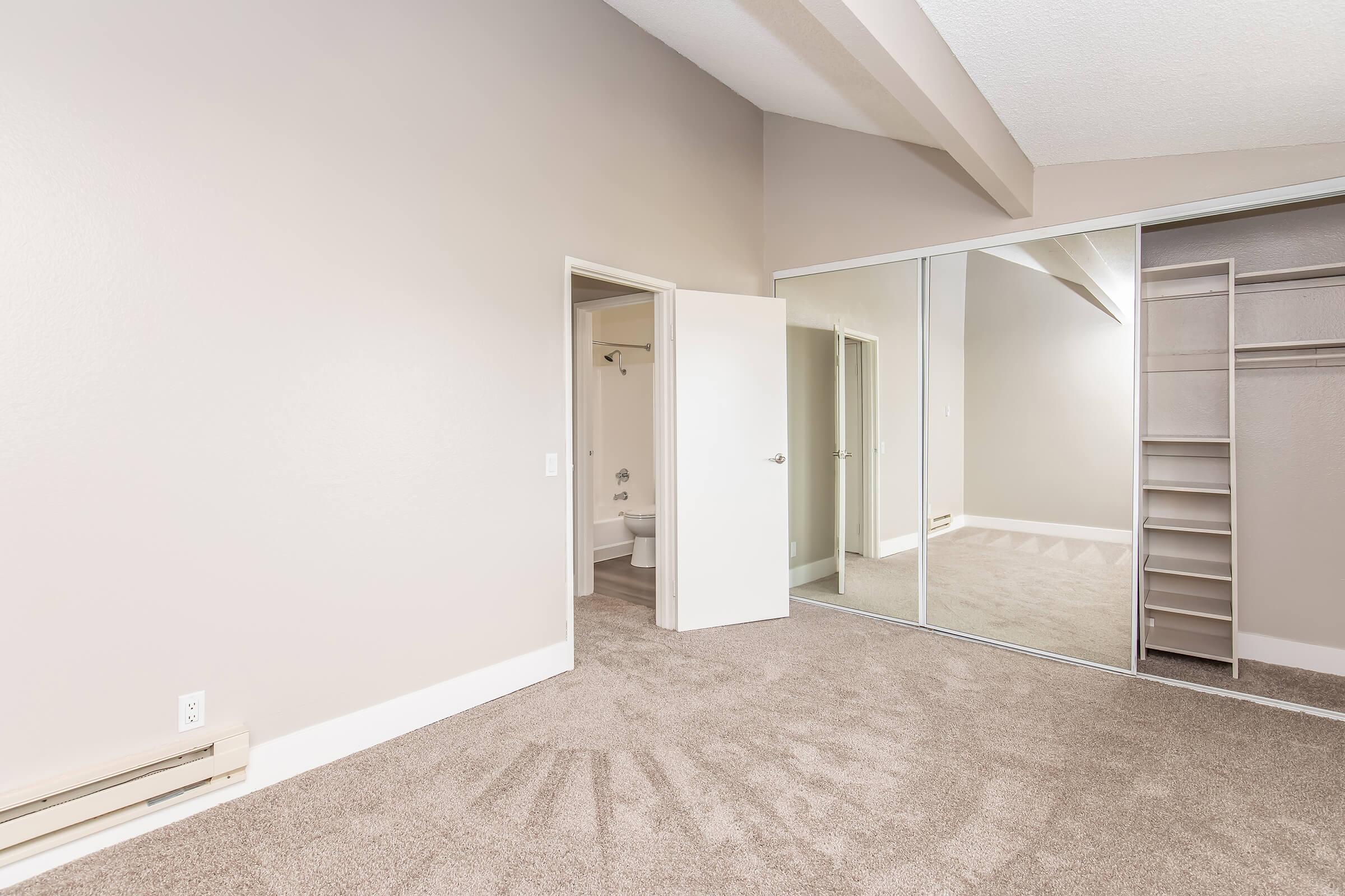 A spacious bedroom with light beige walls and carpet. There are mirrored sliding closet doors on one wall, and a door leading to a bathroom. The room features high ceilings and natural light, creating an inviting atmosphere.
