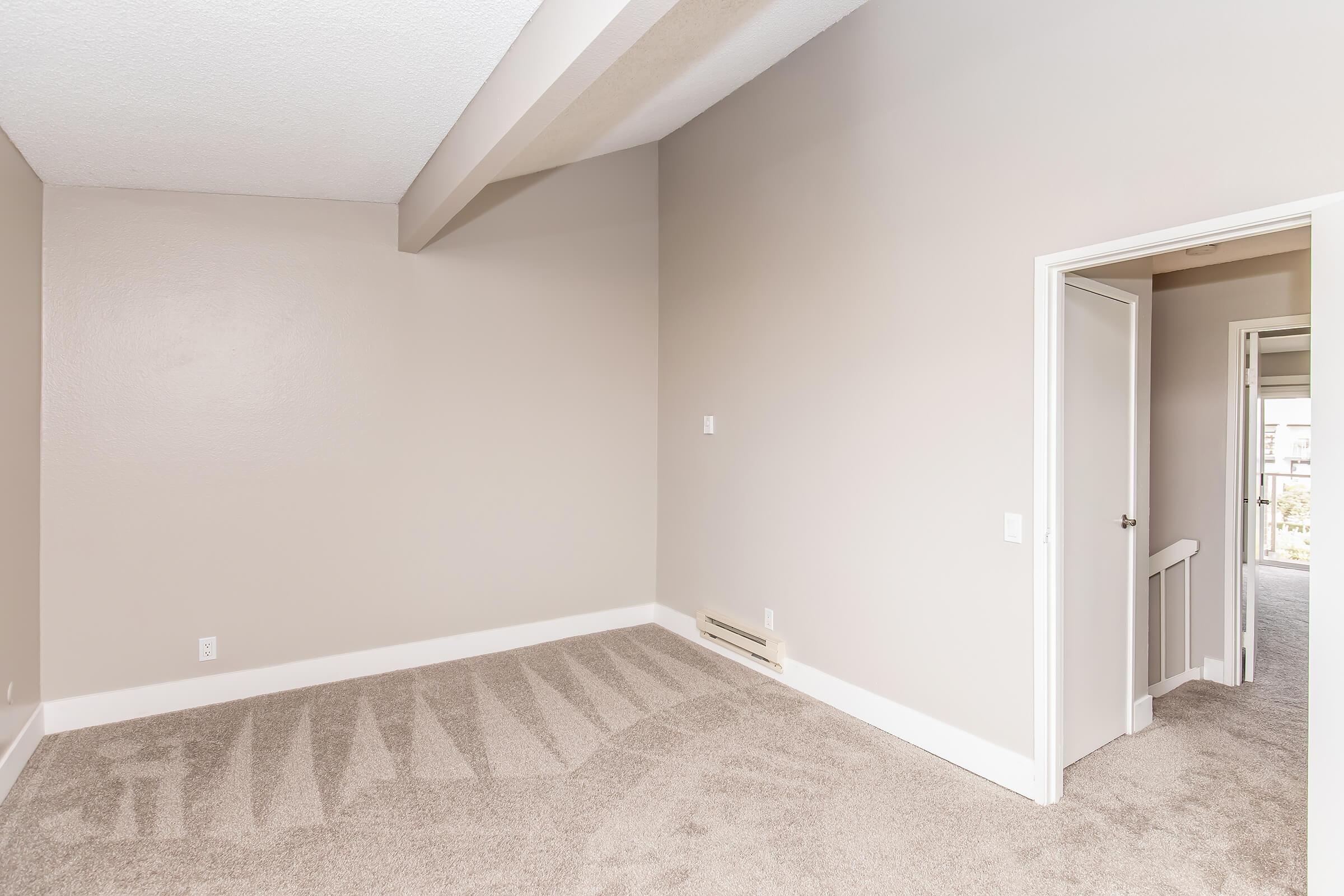 An empty room with light beige walls and a tan carpet. The space features a corner with high ceilings and a doorway leading to an adjacent room. A baseboard heater is visible on the wall. Natural light may enter through the unseen window, creating a bright and open atmosphere.