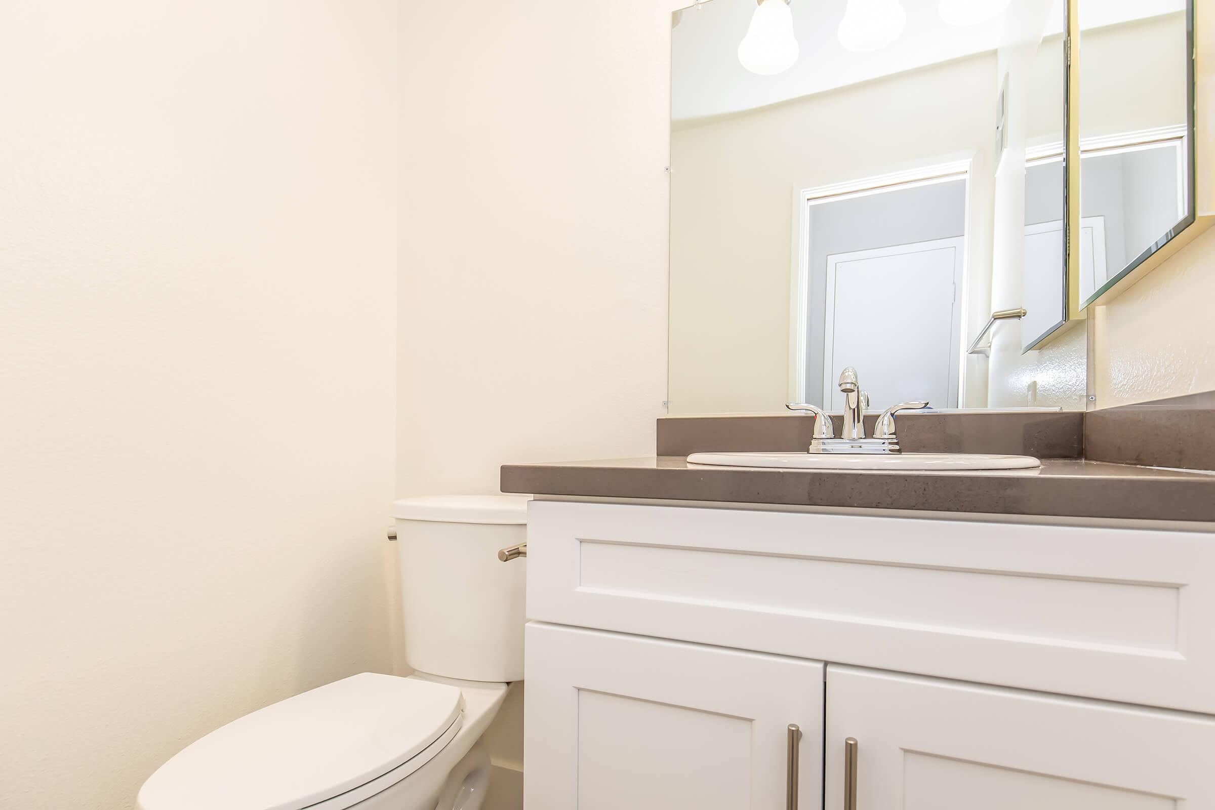 A clean and modern bathroom featuring a white toilet, a sleek vanity with a grey countertop, and a large mirror. Soft natural light illuminates the space, highlighting the minimalist design and neutral color scheme, providing a bright and airy atmosphere.
