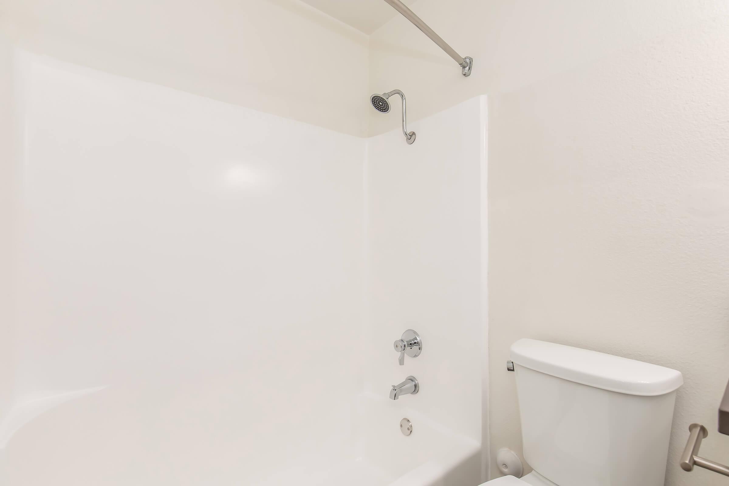 A clean, modern bathroom featuring a white bathtub with a showerhead, a chrome faucet, and a white toilet. The walls are plain and light-colored, creating a bright and spacious atmosphere.