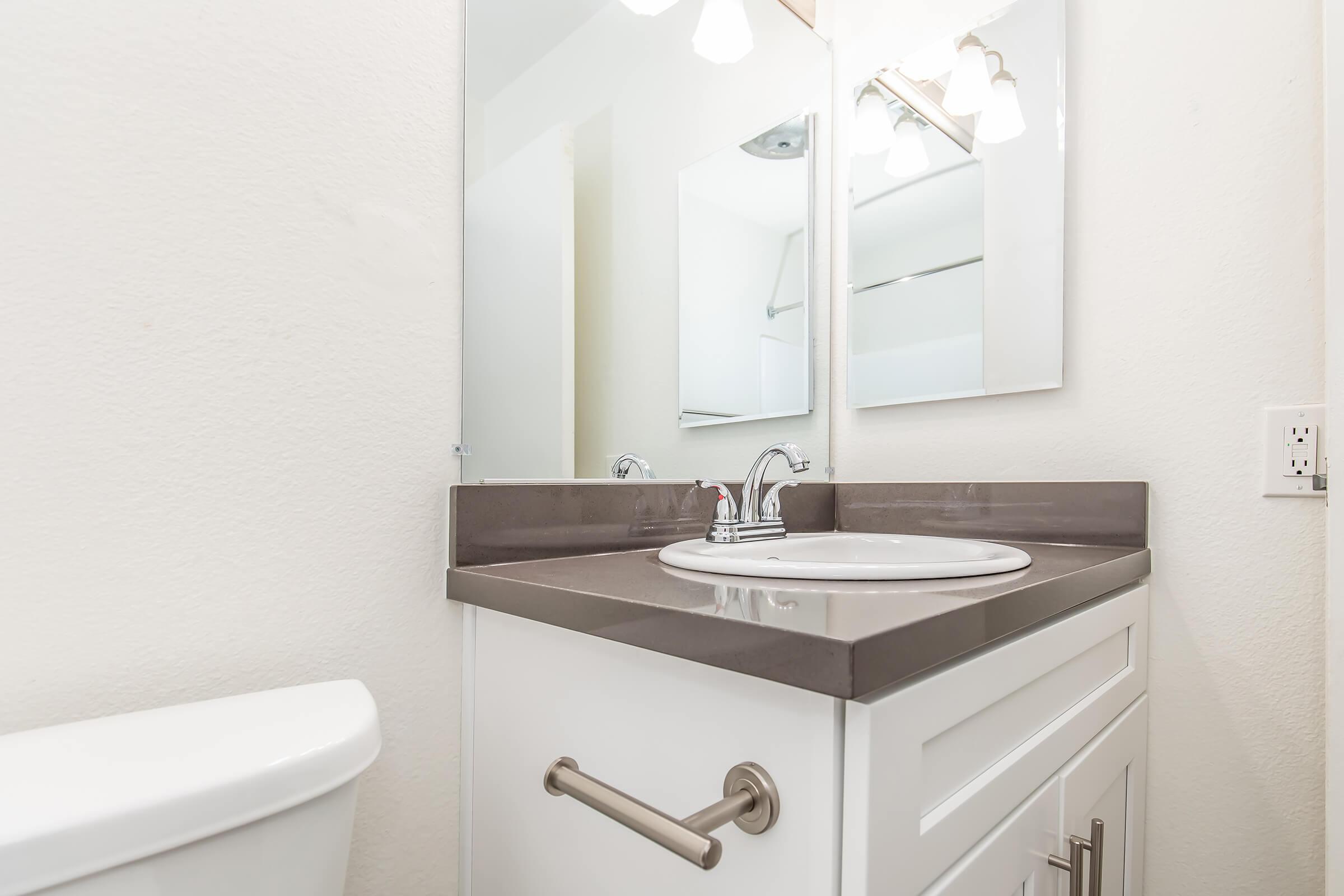 A modern bathroom featuring a white vanity with a round sink and a sleek countertop. There is a large mirror above the sink, and a light fixture with multiple bulbs. A white toilet is visible on the left, and the walls are painted a light color, creating a clean and airy atmosphere.