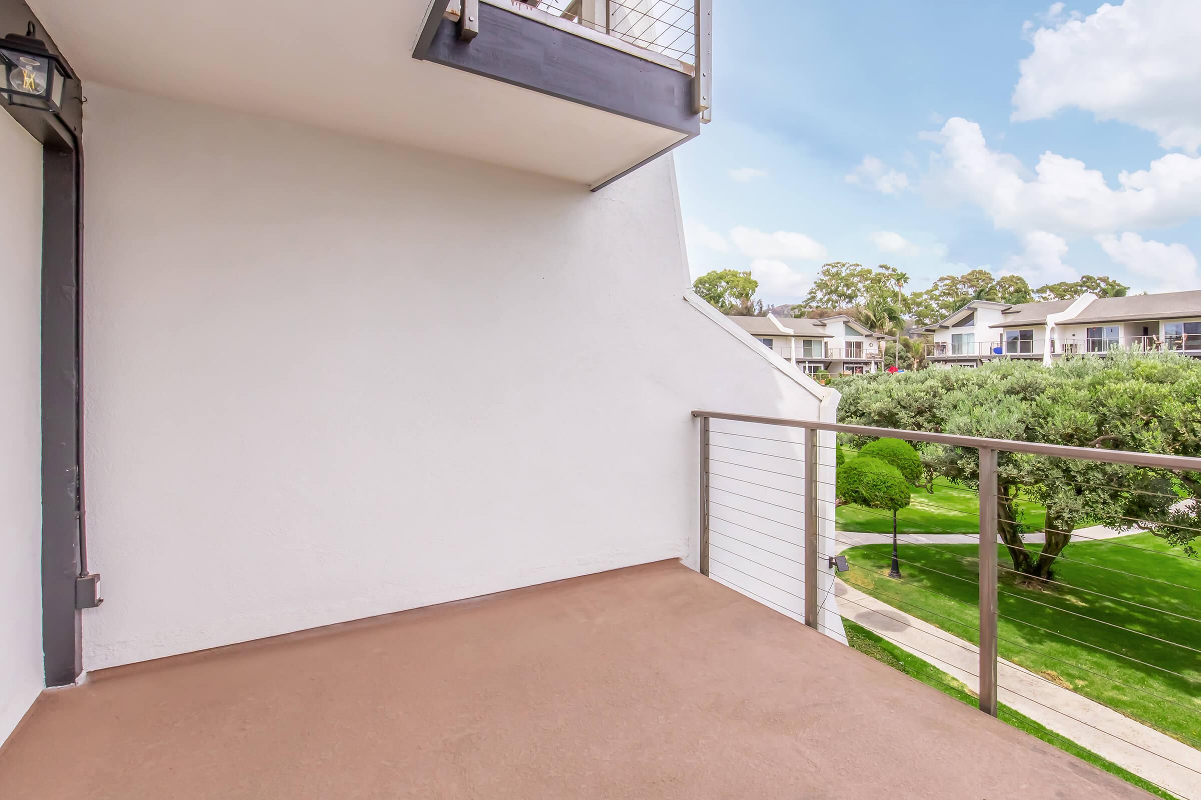 A clean, empty balcony with a modern railing, overlooking a green landscape with neatly trimmed bushes and trees. In the background, there are residential buildings under a bright blue sky with scattered clouds. The scene conveys a peaceful, open outdoor space.