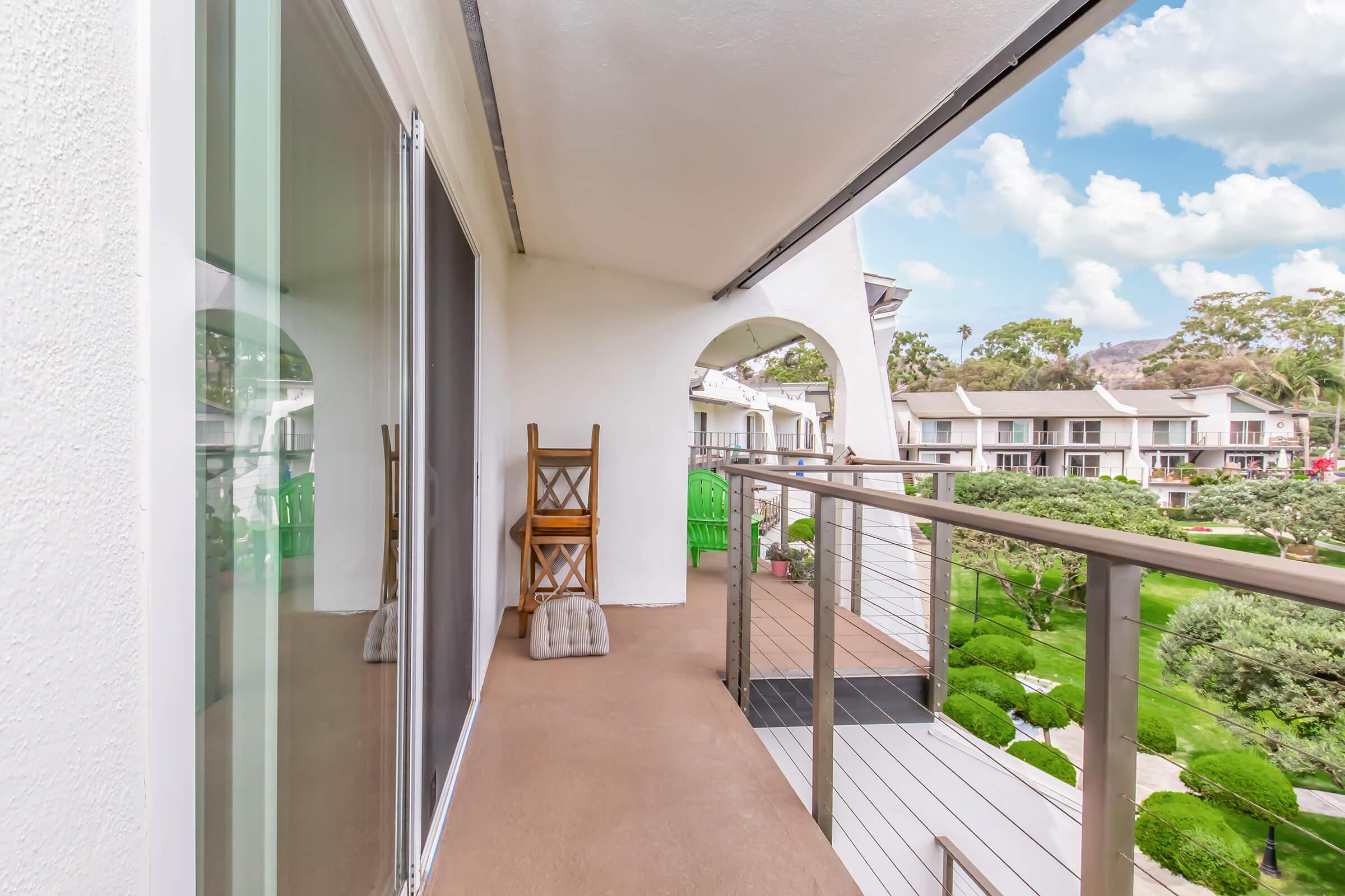 A balcony with a wooden chair and a small table, overlooking a landscaped garden with green shrubs and trees. The view includes a clear blue sky with fluffy clouds, showcasing a peaceful outdoor setting.