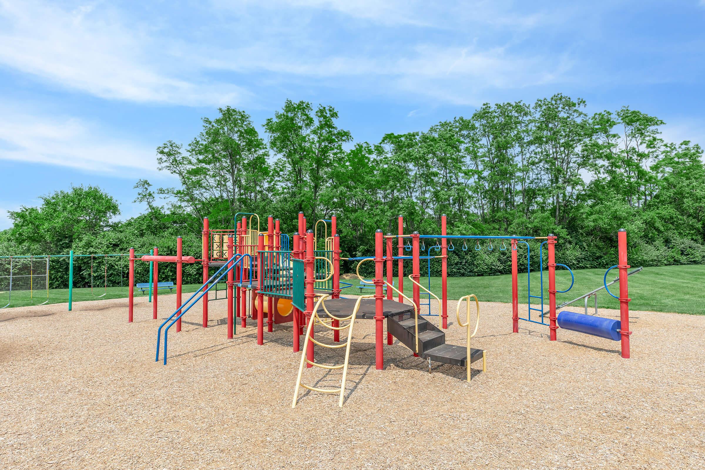 Colorful playground equipment with slides, climbing structures, and monkey bars, set on a tan gravel surface. Lush green trees in the background under a clear blue sky. Ideal for children's outdoor play and activities.