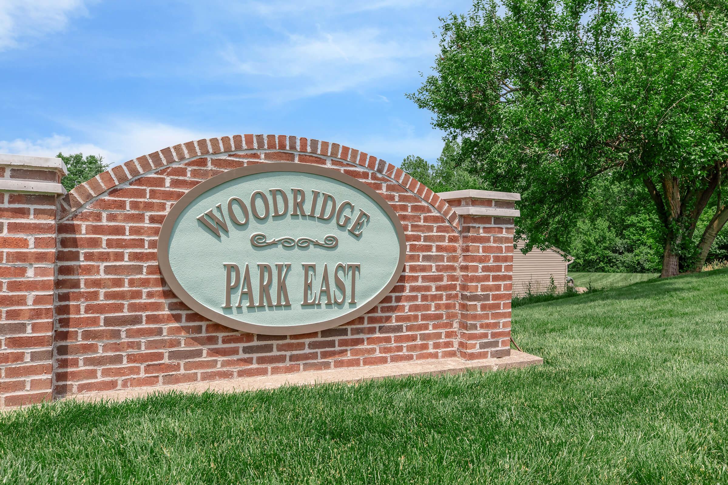 Sign for Woodridge Park East, featuring a curved brick arch design. The sign is set against a backdrop of green grass and trees. The letters are prominently displayed in a decorative font, indicating the name of the park.