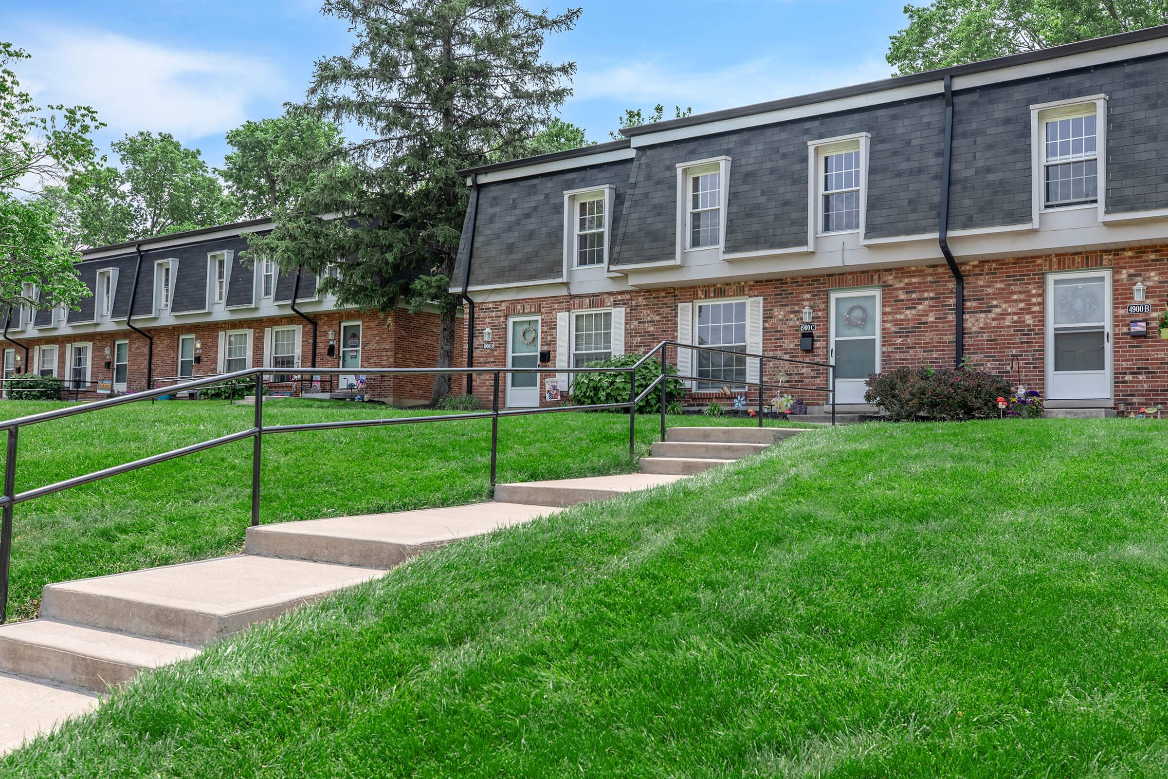 A row of brick townhouses with gray roofs, situated on a green lawn. There are concrete steps leading up to each unit, with a railing for support. Flower beds are visible near the entrances, and trees can be seen in the background under a blue sky.