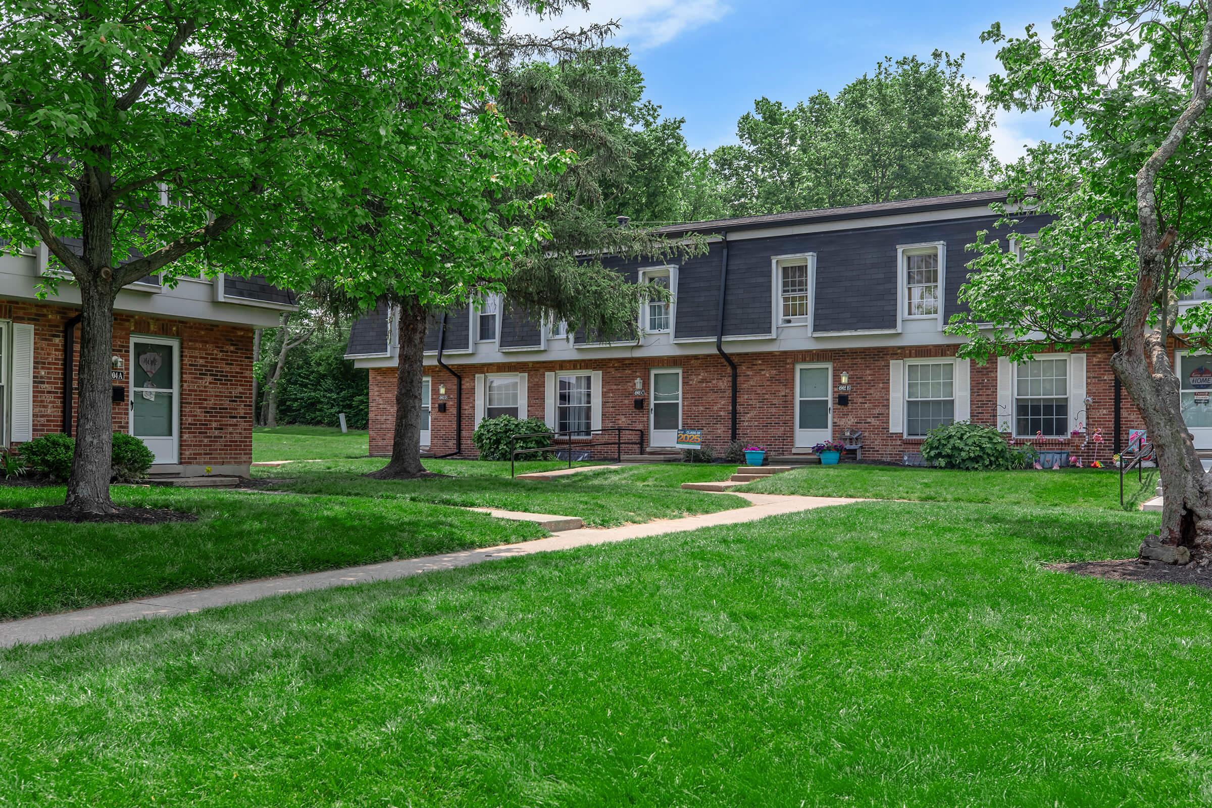 A serene residential area featuring two-story brick apartment buildings surrounded by lush green lawns and trees. The pathway leads through the grass, connecting the buildings, creating a welcoming and peaceful community atmosphere. The sky is slightly cloudy, adding a soft contrast to the greenery.