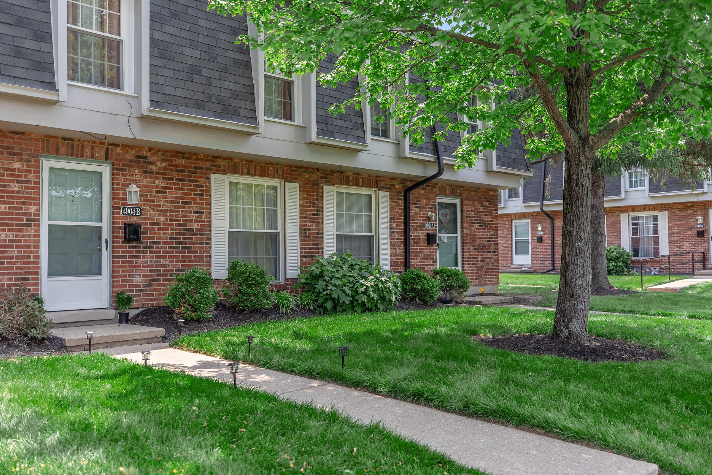 A well-maintained brick townhouse with a gray shingle roof, featuring a green lawn and shrubbery. The walkway leads to two front entrances, surrounded by landscaped grass and small lights. Nearby, another similar townhouse can be seen in the background.