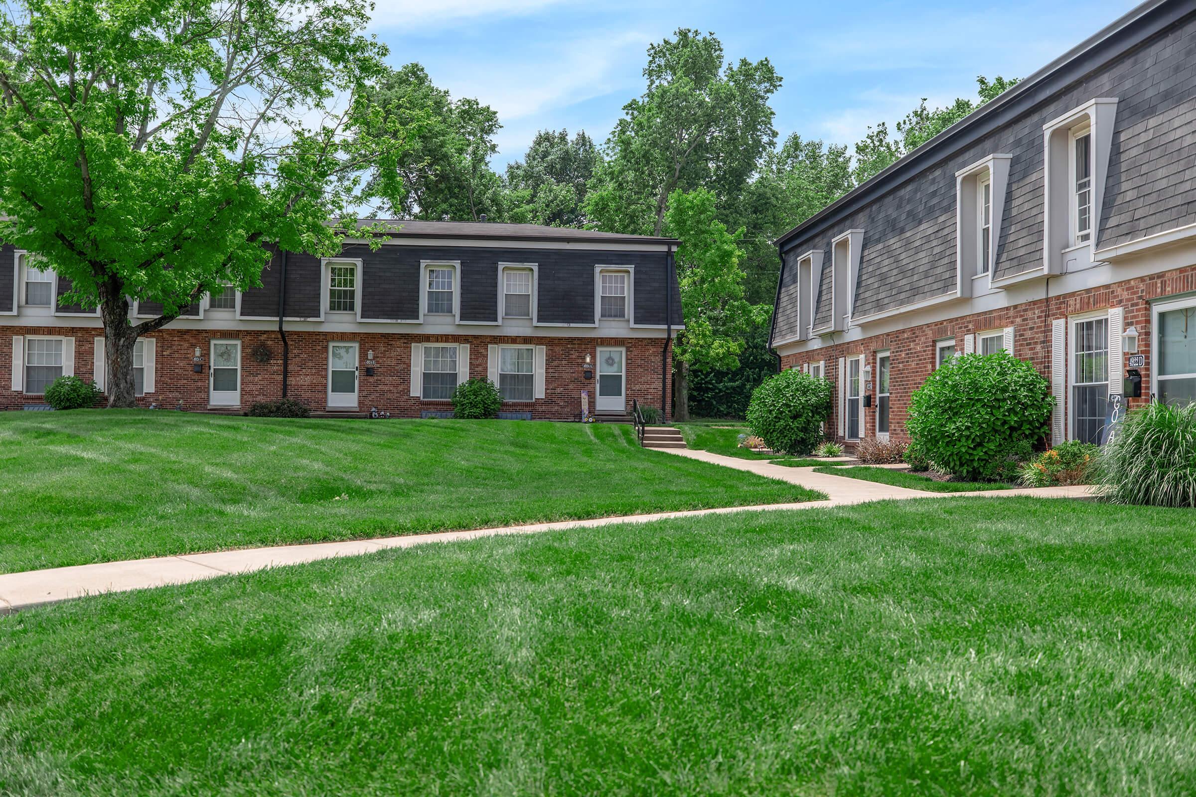A well-maintained residential area featuring two-story brick and siding apartment buildings, surrounded by lush green grass and trees, with a walkable pathway connecting the buildings. Bright blue sky and greenery create a serene atmosphere.