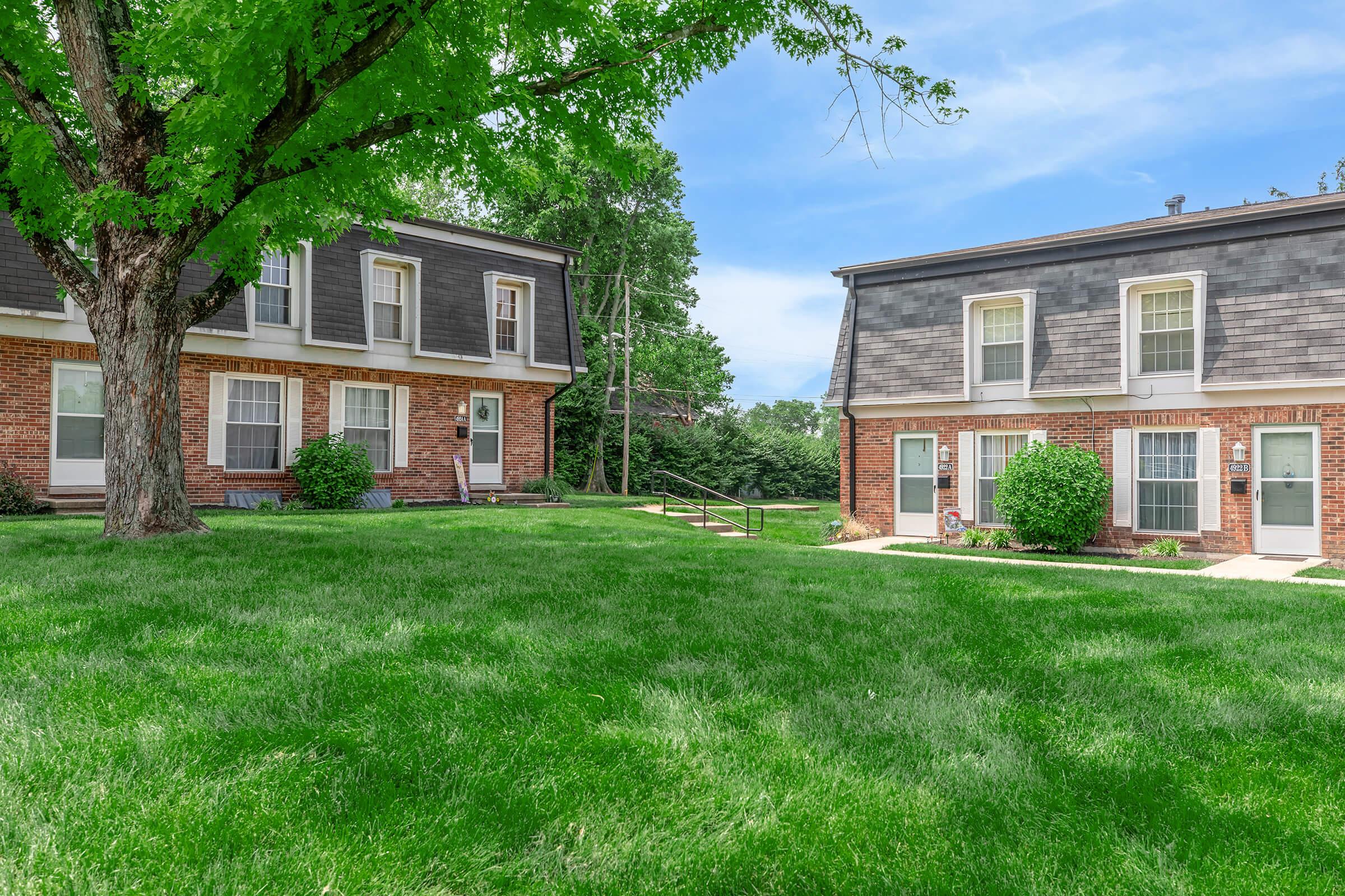 Two two-story brick apartment buildings are situated on a grassy lawn. One building has a porch with steps leading up to the entrance. The area is surrounded by trees and a clear blue sky, creating a peaceful residential environment.