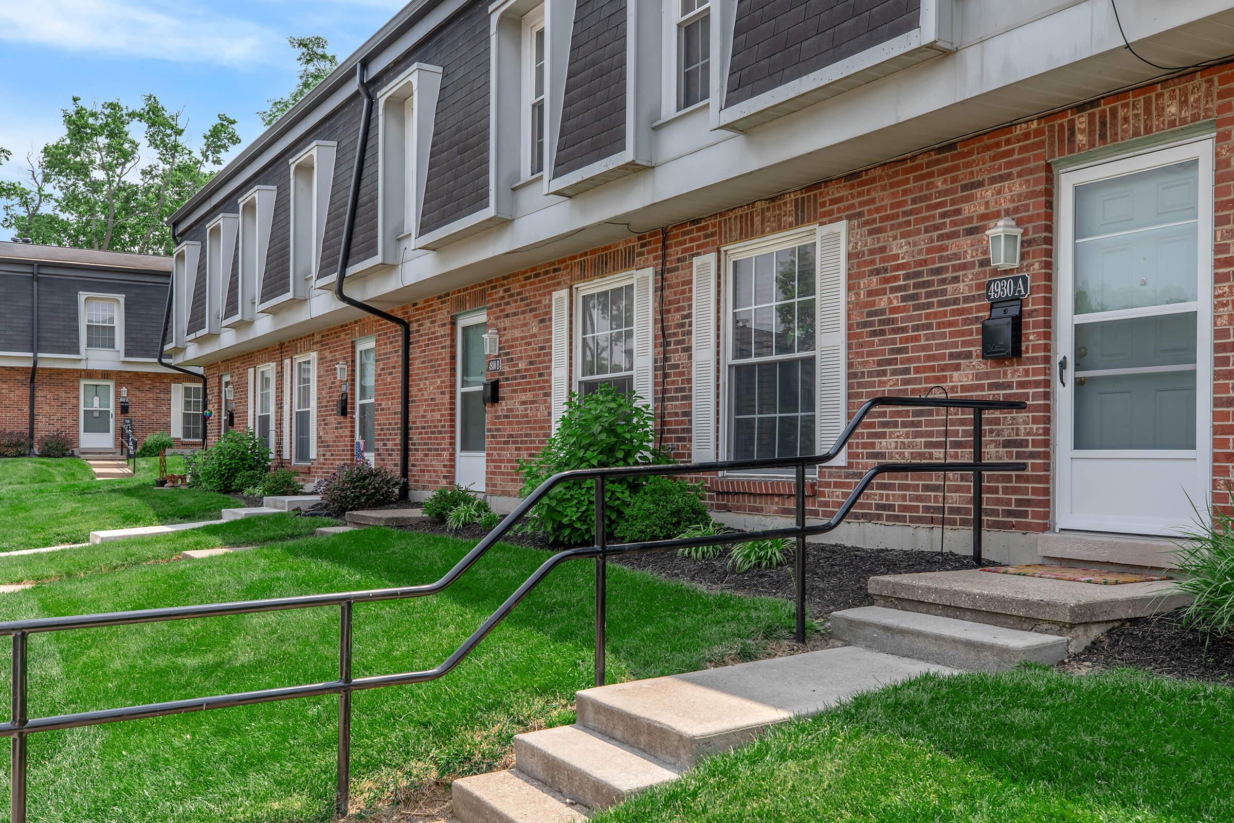 A row of townhouses with brick exteriors, featuring multiple entries. Each unit has a small front step leading to the door, with a lawn area and landscaped greenery in the foreground. The building has a modern design with a mix of brick and siding.