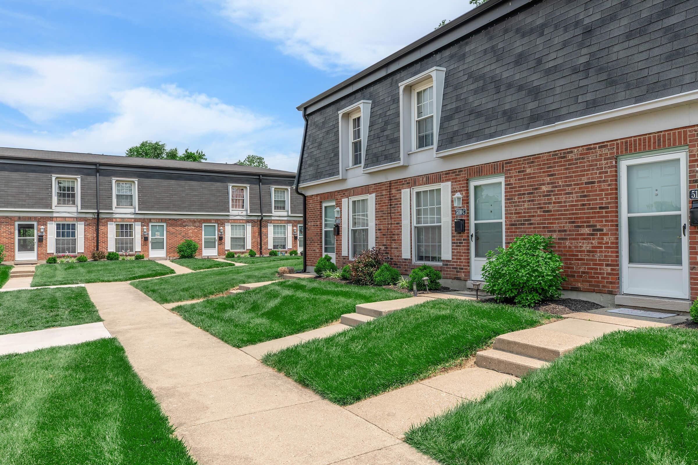 A view of a well-maintained apartment complex featuring brick and gray siding buildings. Sidewalks lead to individual units, surrounded by neatly trimmed green lawns and small shrubs. The sky is clear with a few clouds, creating a bright and inviting atmosphere.