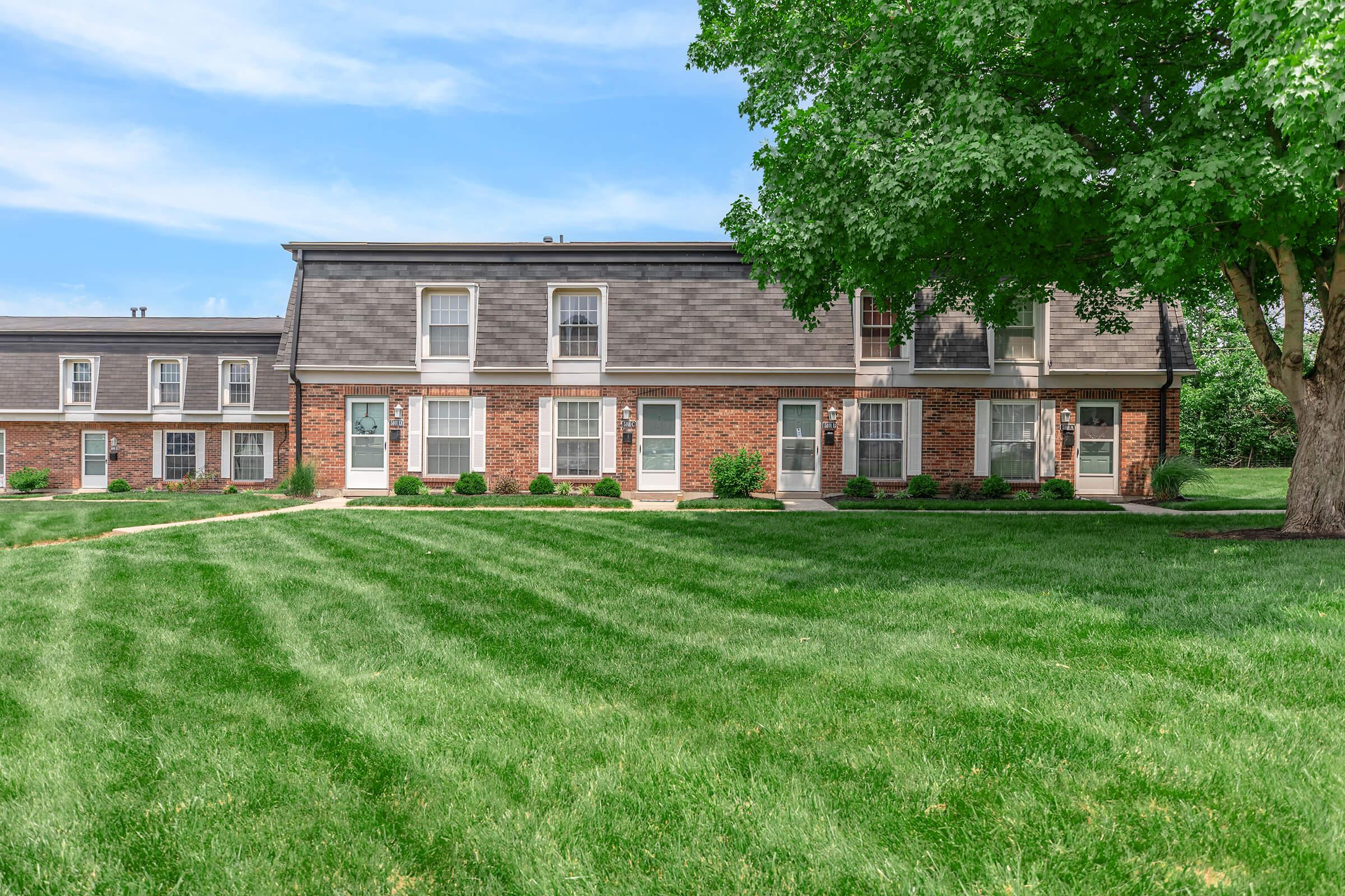 A well-maintained brick apartment building with multiple units, surrounded by lush green grass and landscaped bushes. The sky is clear with a few clouds, and a large leafy tree is visible on one side.