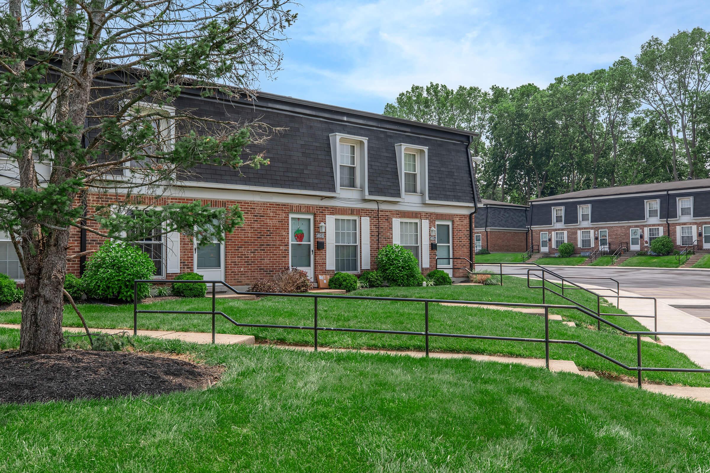 A residential area featuring two-story brick apartment buildings with dark roofs, surrounded by well-maintained green lawns and shrubs. A concrete pathway with handrails leads to the entrances, while trees and a clear blue sky provide a pleasant backdrop.
