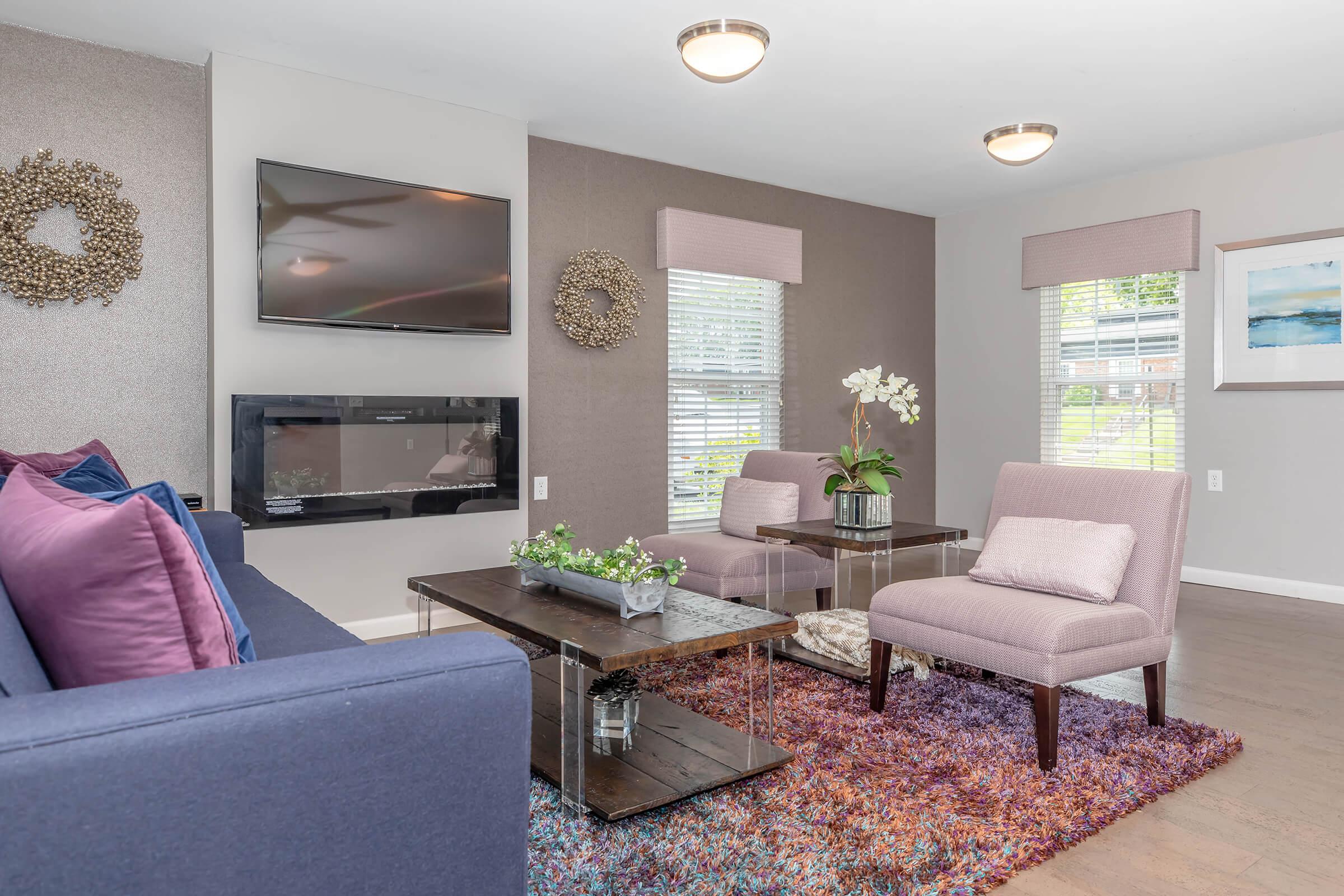 Cozy living room featuring a blue sofa with purple and pink cushions, two light pink chairs, a coffee table with a plant, and a flat-screen TV mounted on the wall. Natural light filters through window shades, and decorative wall accents add a touch of elegance to the space.