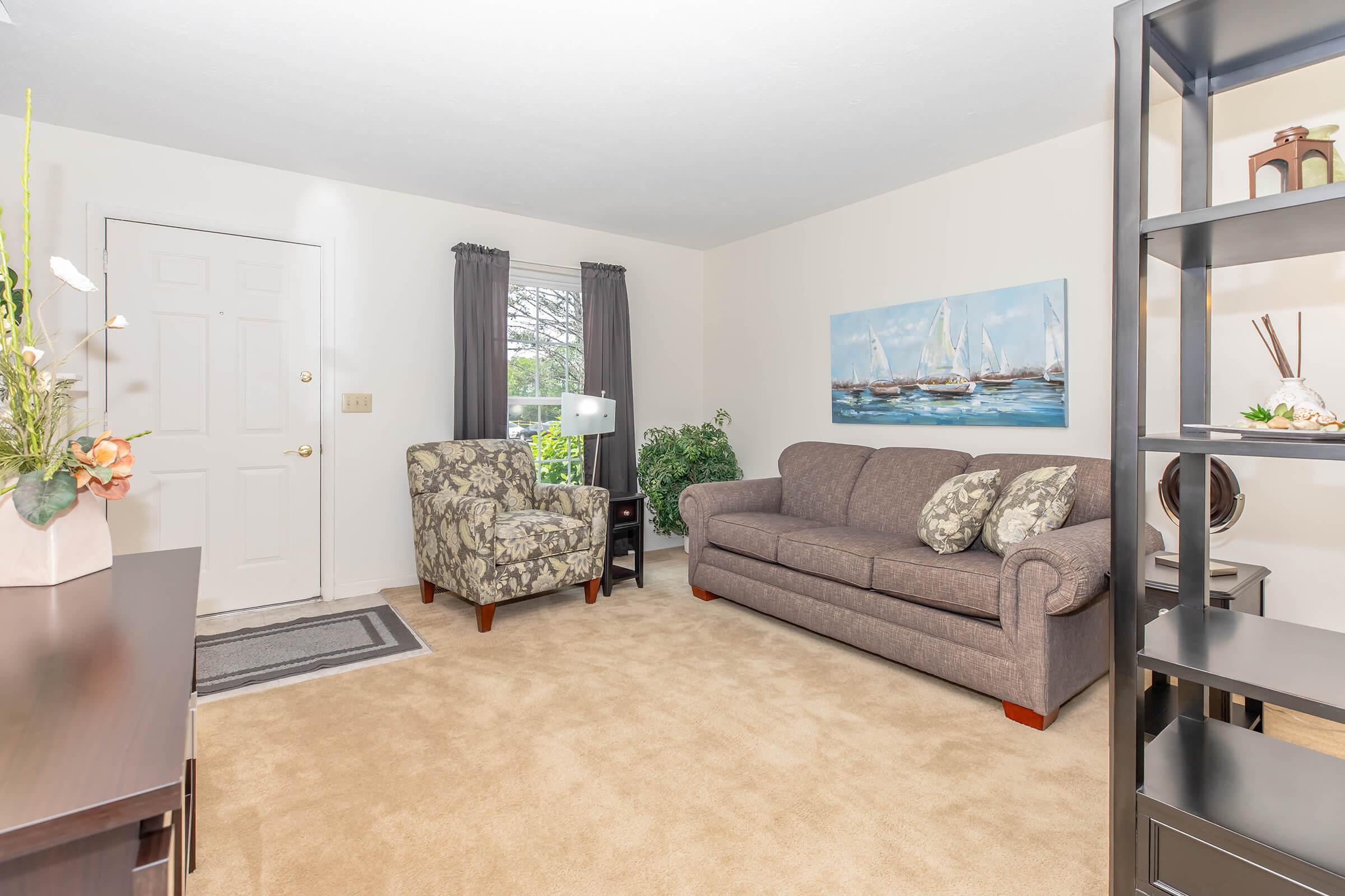 Cozy living room featuring a beige carpet, a gray sofa, and a patterned armchair. A painting of sailboats hangs on the wall, complemented by indoor plants. Large windows let in natural light, and a decorative shelf adds to the room's charm, creating an inviting atmosphere.