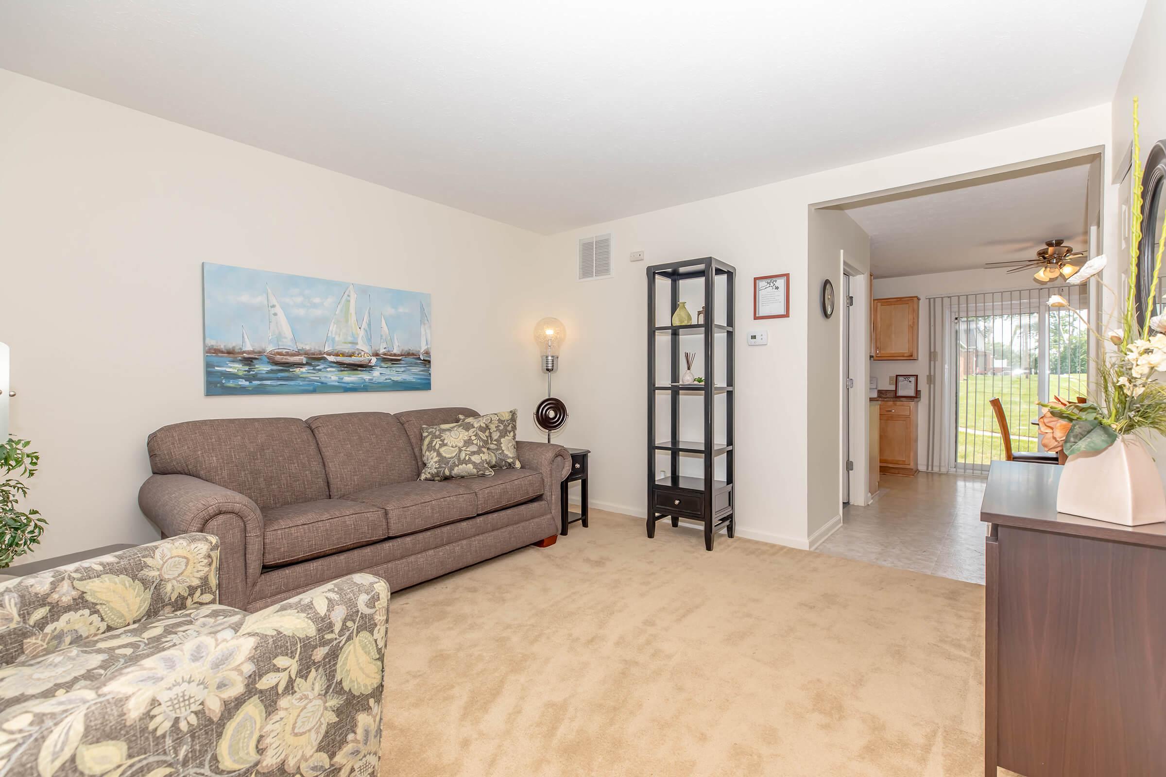 Cozy living room featuring a brown sofa with decorative pillows, a floral patterned chair, and a tall black shelving unit. A nautical-themed painting adorns the wall. Natural light streams through a sliding door leading to an outdoor area, creating a warm and inviting atmosphere.