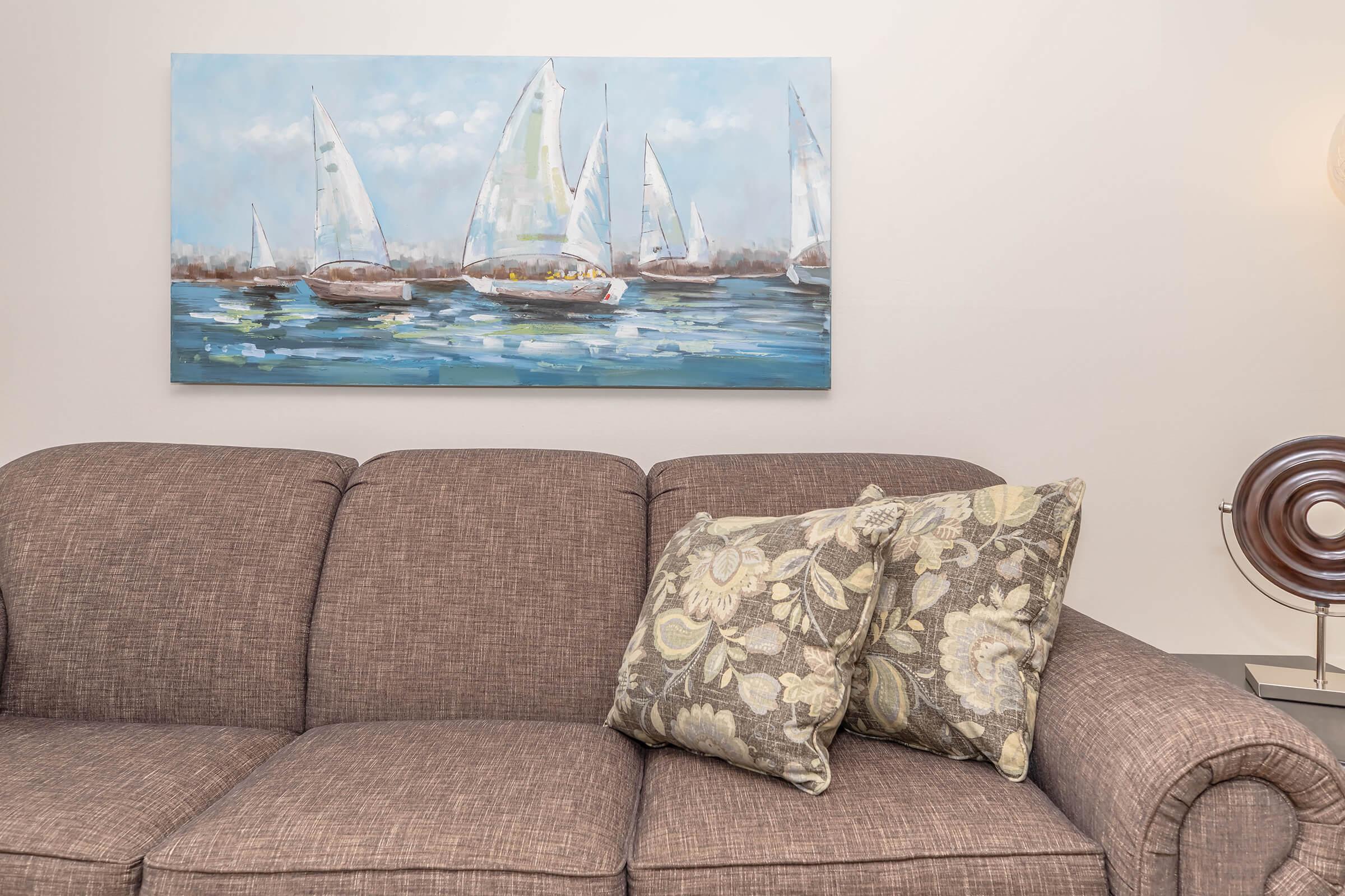 A cozy living room scene featuring a brown couch with floral-patterned pillows. Above the couch hangs a painting of sailboats on a blue sea, adding a nautical touch to the decor. A modern lamp and decorative object are subtly placed in the background, enhancing the inviting atmosphere.