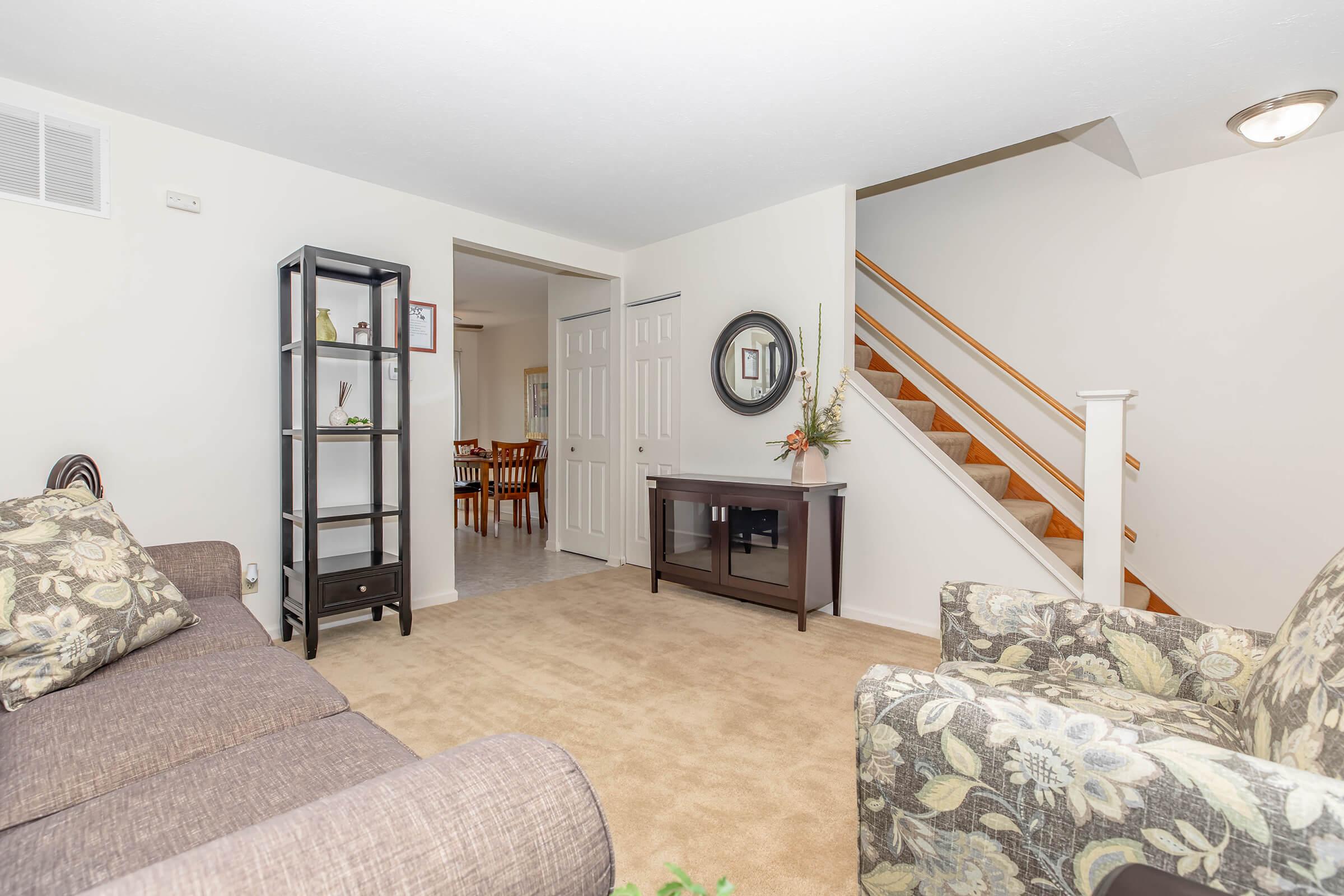 A cozy living room featuring a brown sofa with floral patterned chairs, a small black bookshelf, and a dark wooden TV stand. The room has beige carpet and light-colored walls, with a view of a staircase leading to the upper level and a doorway leading to a dining area in the background.