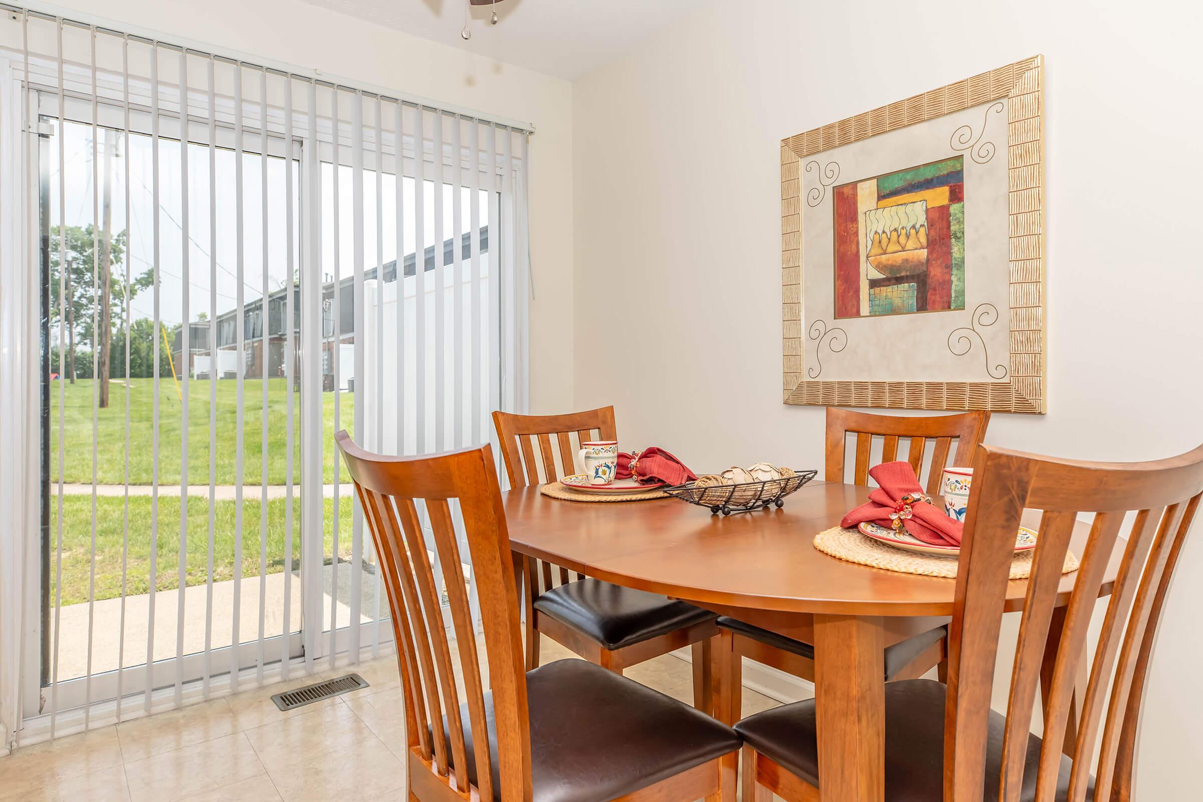 A cozy dining area featuring a wooden table with four chairs arranged around it. The table is set with plates and napkins. A decorative wall hanging adds a touch of color. Large sliding glass doors with vertical blinds open to a view of greenery outside.