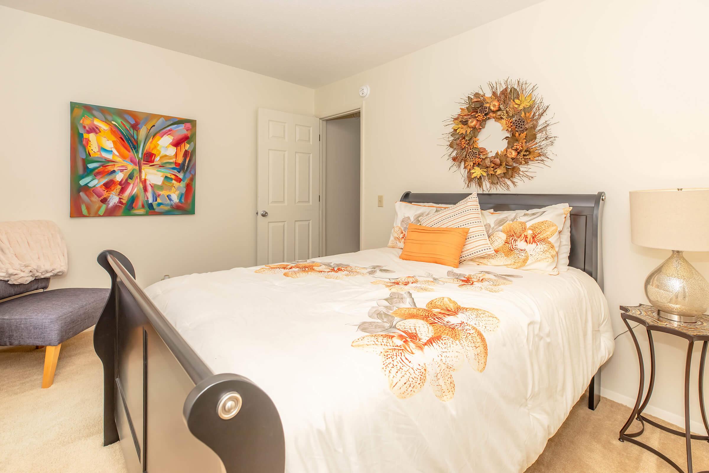 A cozy bedroom featuring a wooden bed with a floral-patterned comforter, an orange accent pillow, a decorative wreath on the wall, and a colorful abstract painting. A nightstand with a lamp and a gray armchair are also visible, creating a warm and inviting atmosphere.