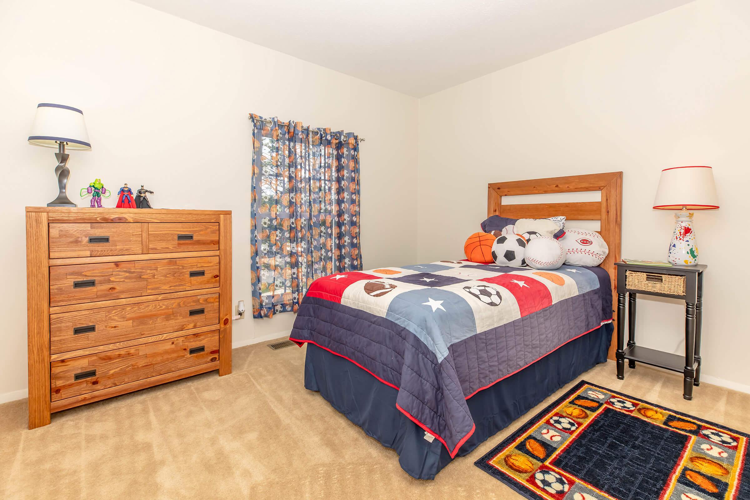 A cozy child's bedroom featuring a wooden bed with a sports-themed quilt, plush toys including a soccer ball and basketball. There's a wooden dresser and a small black nightstand with a lamp. Colorful curtains adorn the window, complementing a fun rug with a sports design on the floor.