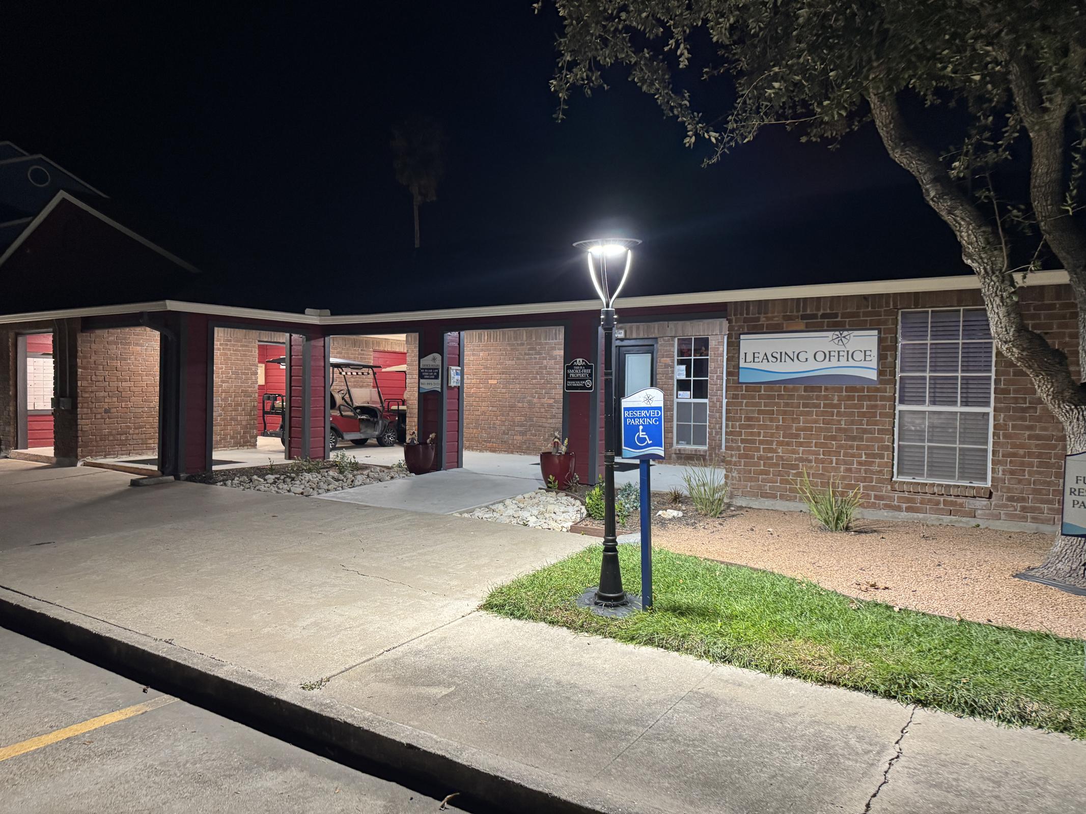 A nighttime view of a leasing office building with brick walls. A lamp post illuminates the entrance, which features a sign indicating "Leasing Office." The parking area is visible, along with a small patch of grass and shrubs. There's a service vehicle partially visible inside the office.