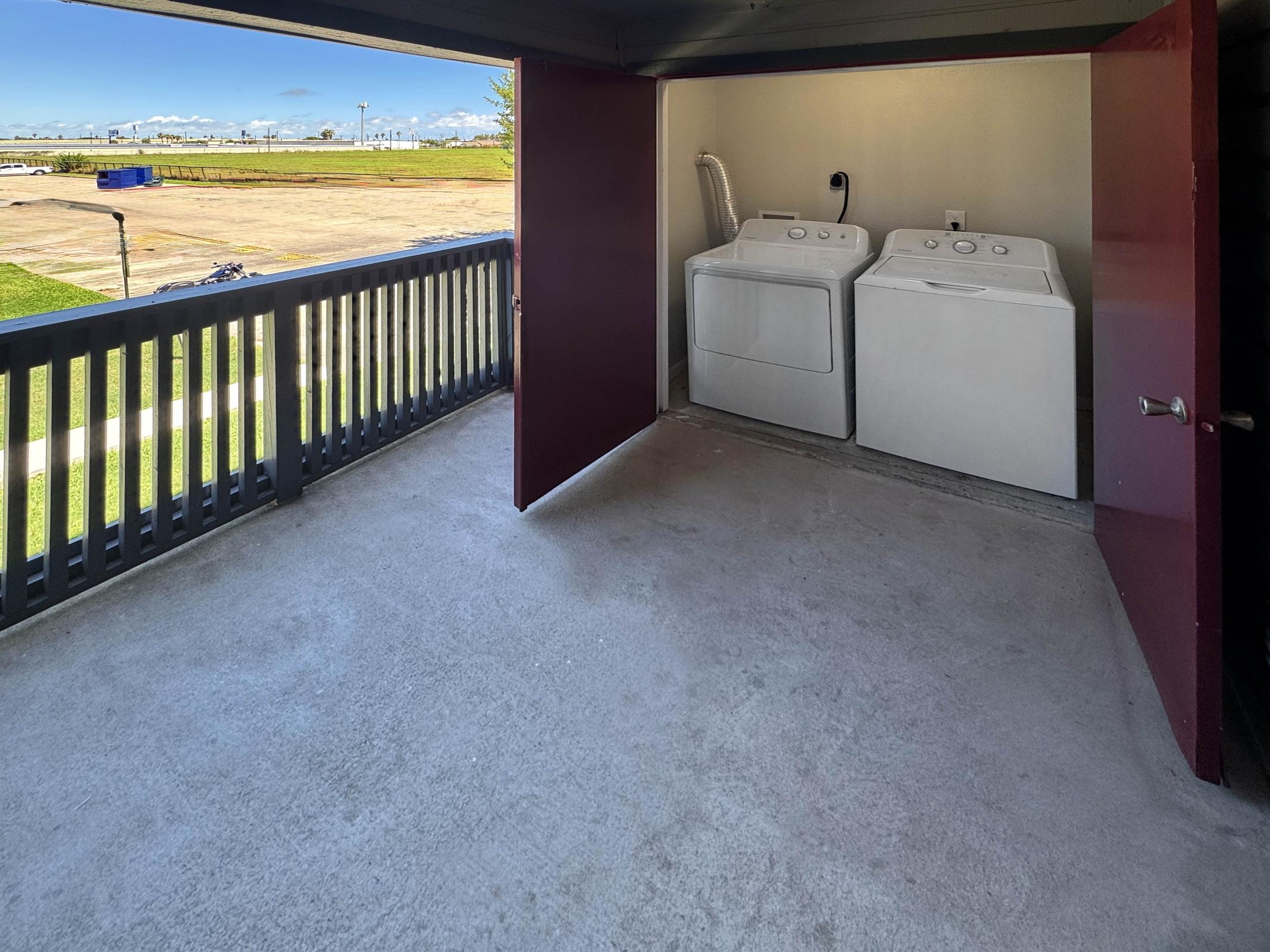 A laundry area featuring two white washing machines placed side by side inside a small enclosed space. The open door reveals a clean, empty concrete floor and a view of green grass and sky outside.