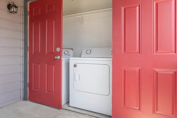 A laundry room inside a home, featuring a set of white washing machines behind bright red double doors. The area is well-lit and organized, with a neatly maintained indoor space visible.