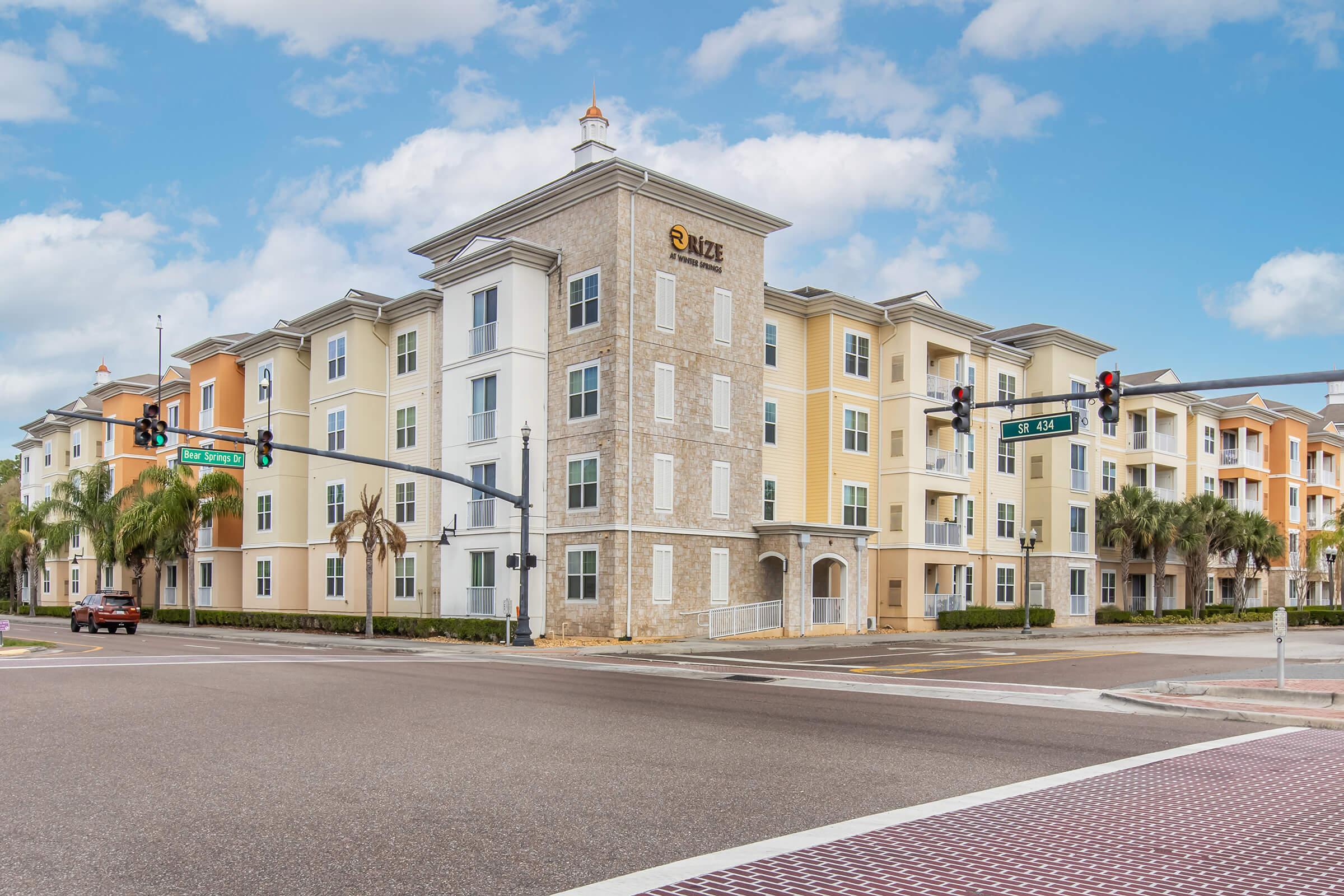 A modern apartment building with a light stone exterior and colorful siding, located at a street corner. Palm trees are visible in the foreground, and traffic lights hang above the intersection. The sky is bright with scattered clouds, creating a welcoming atmosphere.
