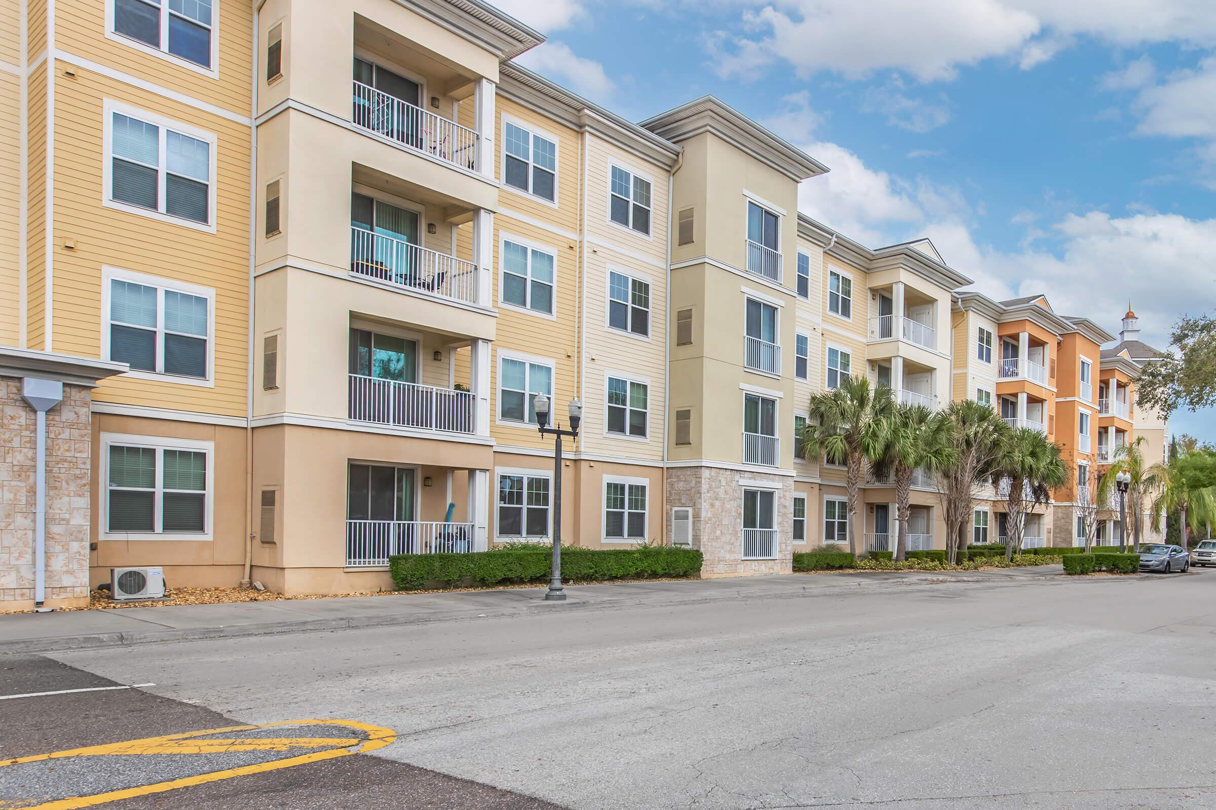 A row of modern, multi-story apartment buildings with varying pastel colors, featuring balconies and large windows. Palm trees are planted alongside the buildings, and a street runs in front of them. The sky is partly cloudy, suggesting a bright day.