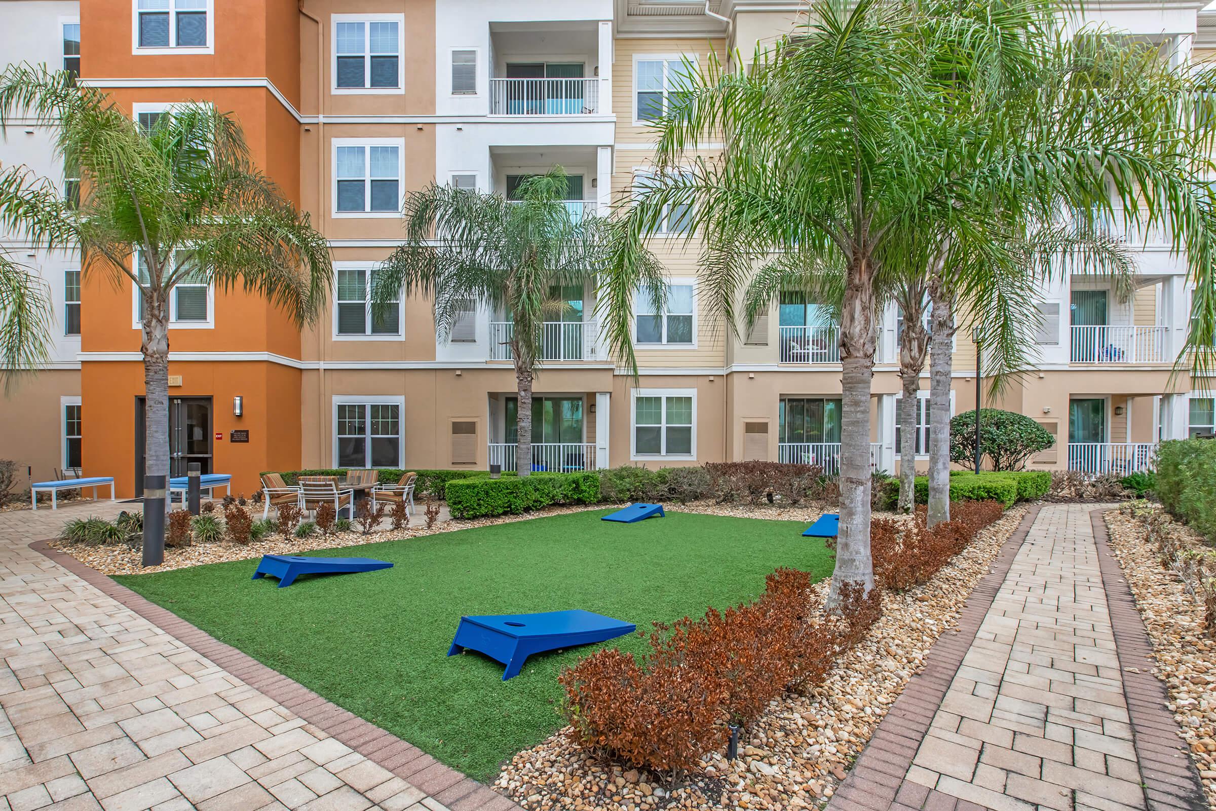 A landscaped courtyard area featuring green artificial grass, palm trees, and blue cornhole boards. Surrounding the space are well-maintained walkways and colorful shrubbery, set against the backdrop of a modern multi-story apartment building with balconies.