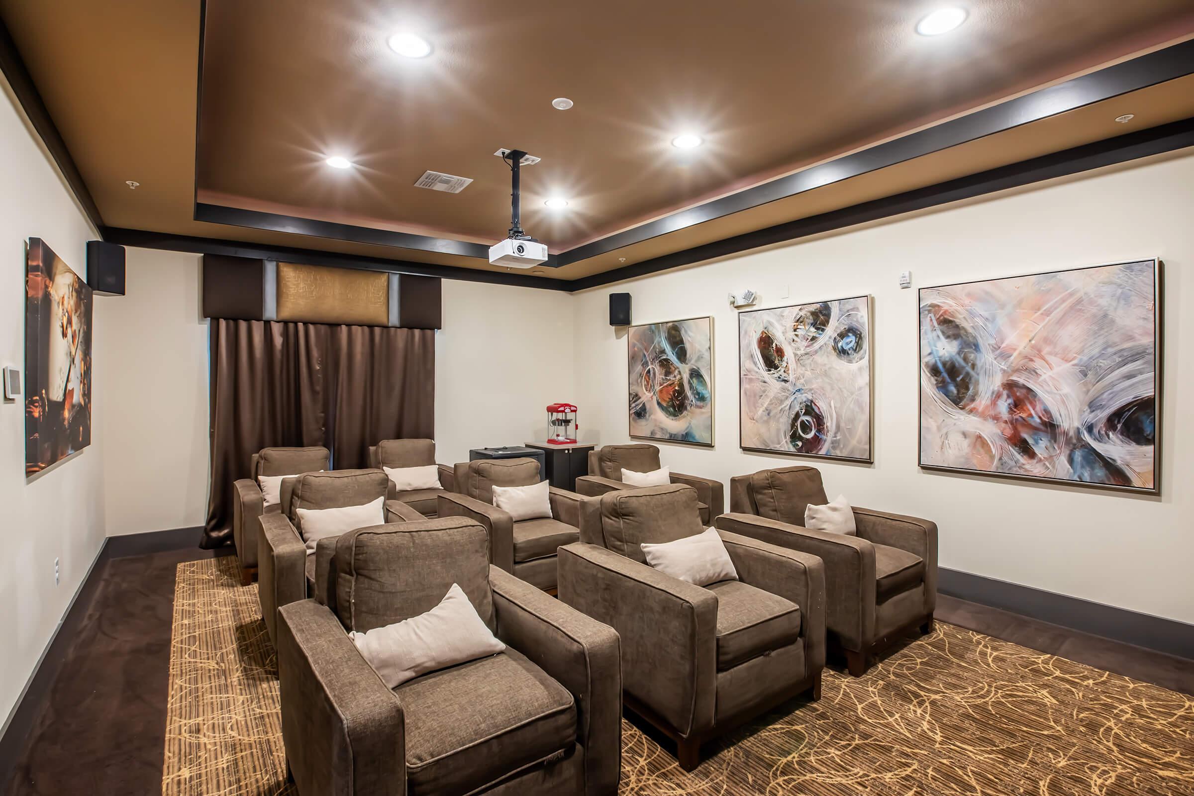 A cozy home theater room featuring multiple comfortable armchairs arranged in rows. The space is illuminated by soft ceiling lights and has a projector for viewing. Two abstract artworks adorn the walls, while dark curtains frame the window. A small table with popcorn adds a casual touch.