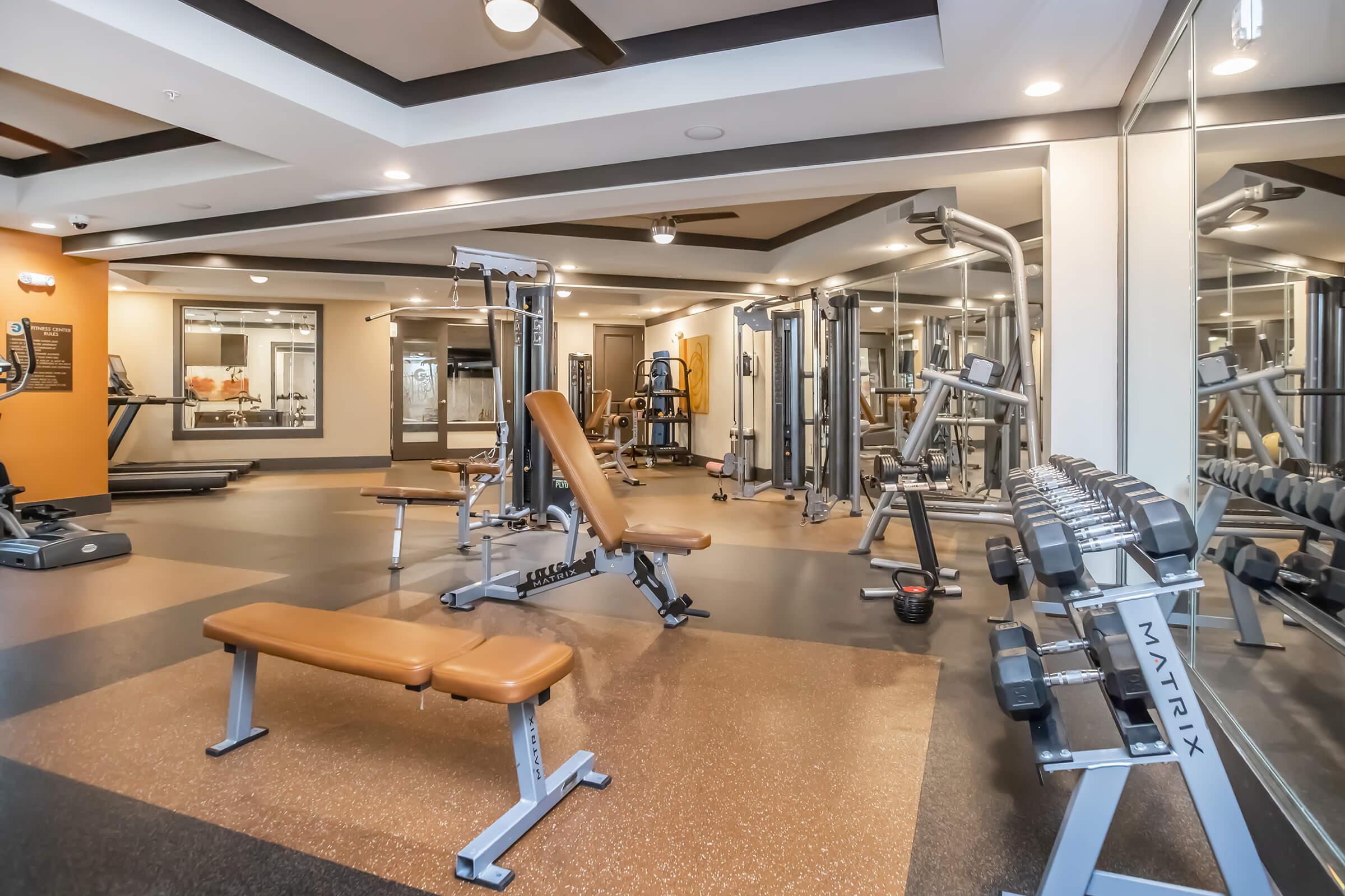 A modern gym interior featuring workout equipment including a weight bench, free weights, and various exercise machines. The space is well-lit with a sleek design, showcasing a blend of dark and light flooring. Mirrors are visible on the walls, enhancing the spacious feel of the room.