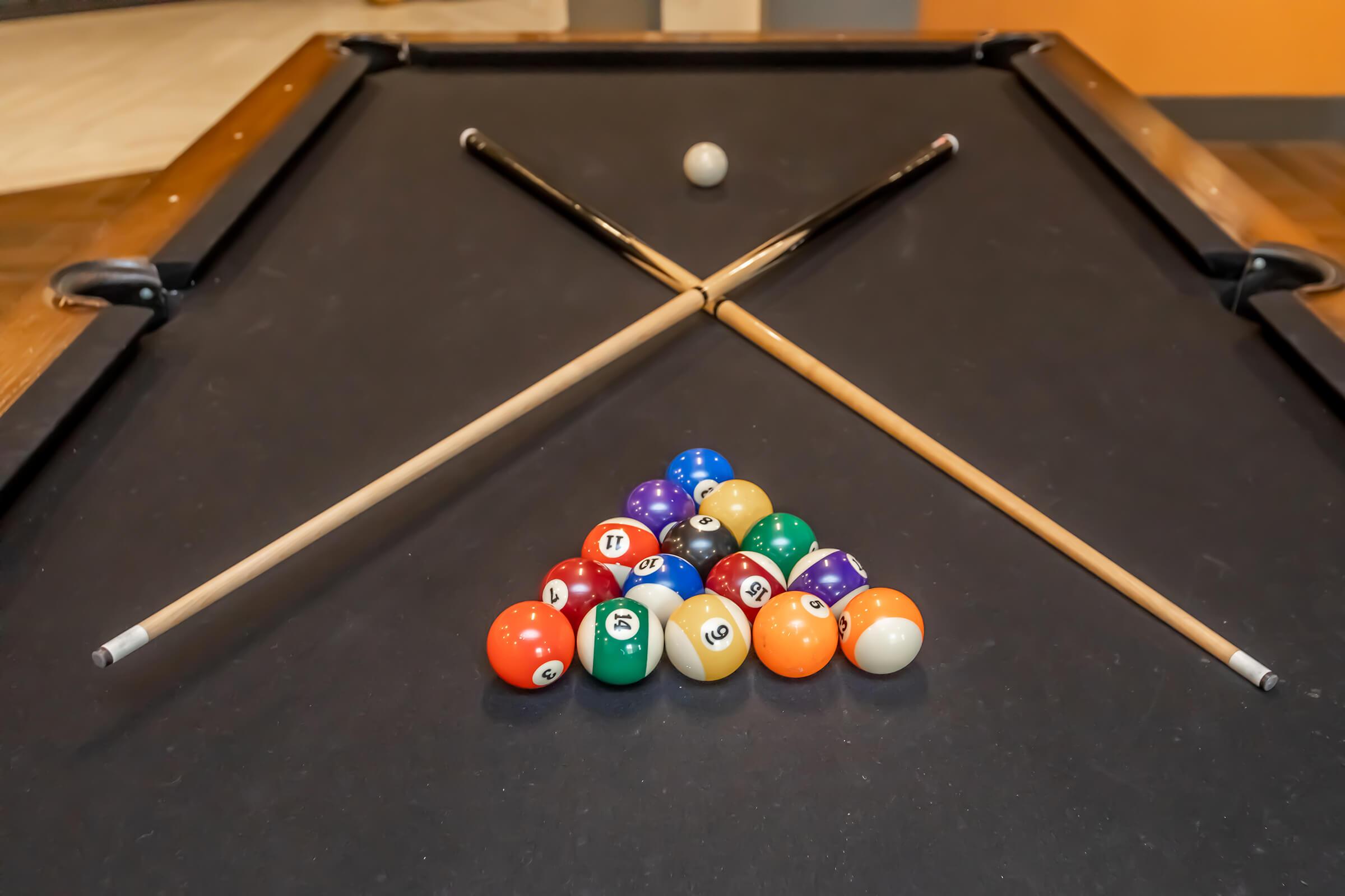 Two pool cues crossed over a triangular arrangement of billiard balls on a slate pool table, with a single cue ball positioned above the triangle. The balls are in various colors, with some numbered. The background features muted tones, creating a casual gaming atmosphere.