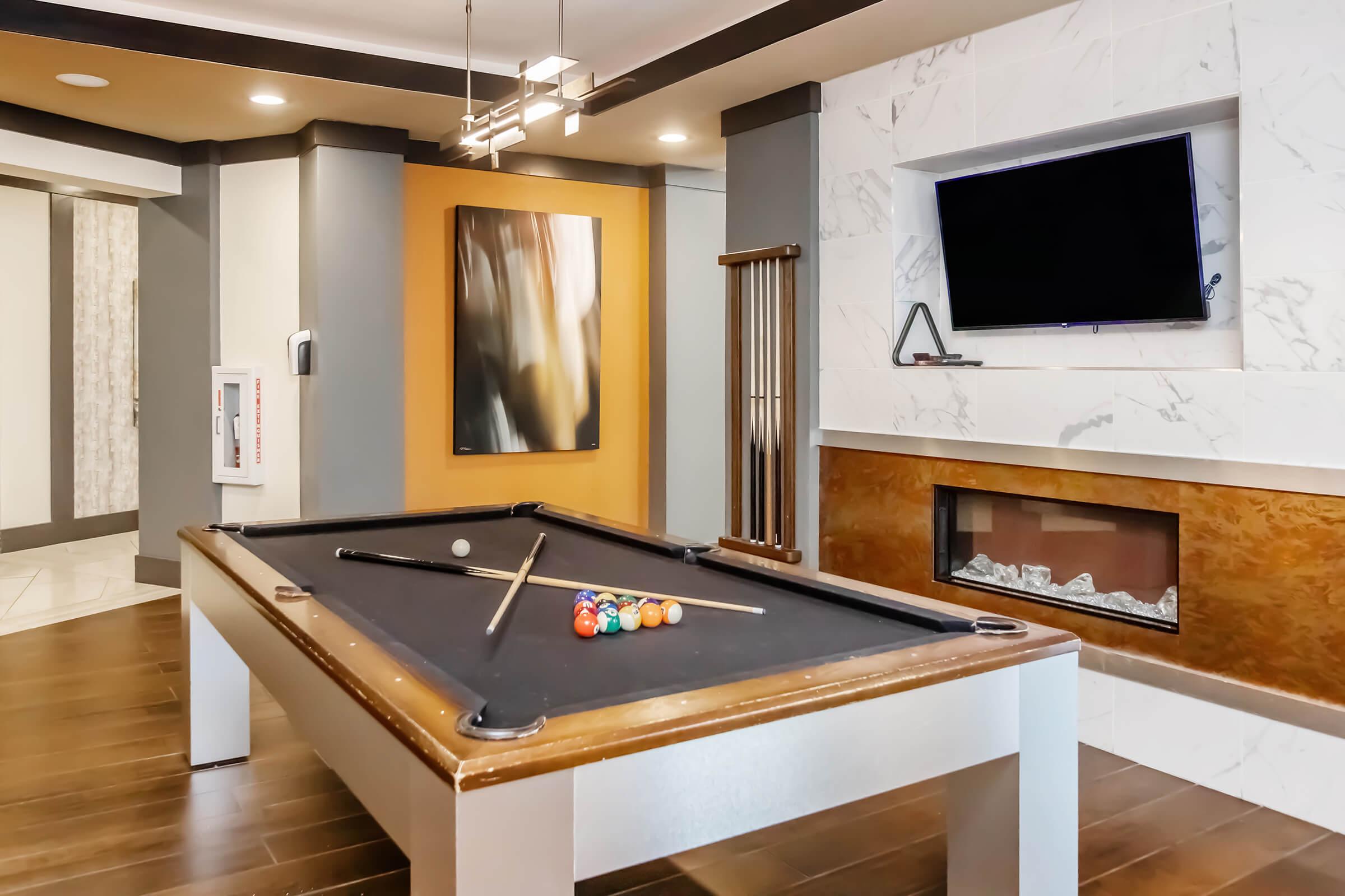 Interior view of a modern game room featuring a sleek pool table with colorful balls arranged on it. The room has a stylish design with warm wood tones, a contemporary fireplace, an abstract painting on the wall, and a mounted flat-screen TV overhead. Soft lighting adds a cozy ambiance.