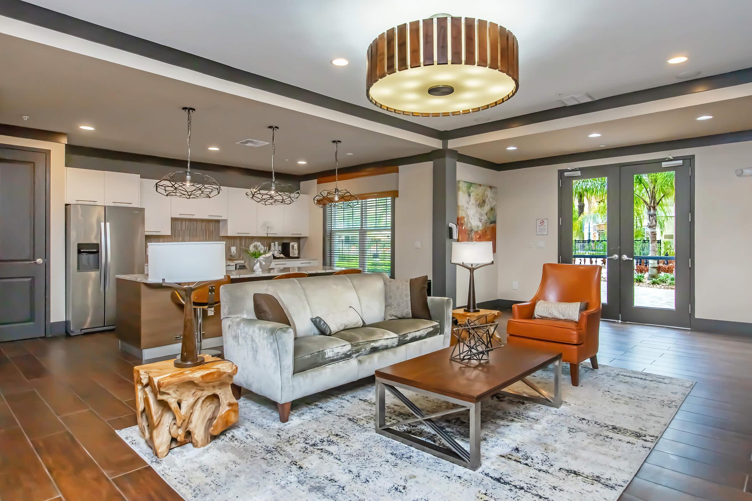 A modern living room featuring a light gray sofa, an orange armchair, and a wooden coffee table on a patterned rug. Pendant lights hang from the ceiling, and large windows allow natural light in. The kitchen is visible in the background, with sleek appliances and a clean design.