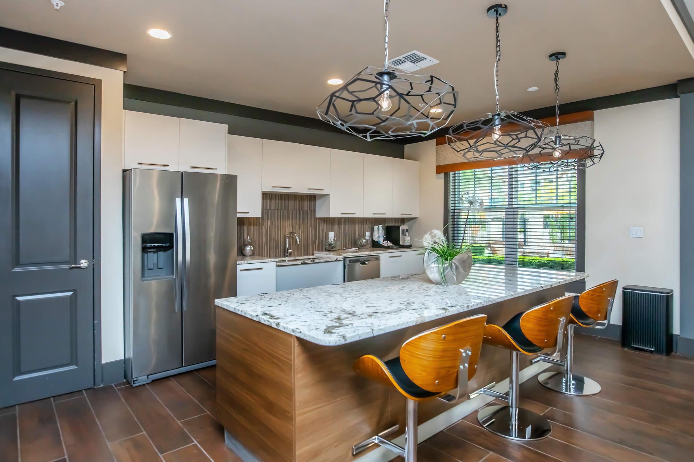 Modern kitchen with sleek white cabinets and a granite countertop island featuring three wooden bar stools. Stainless steel refrigerator and appliances visible, along with decorative vases. Large windows provide natural light, and unique pendant lights hang from the ceiling, enhancing the contemporary design.