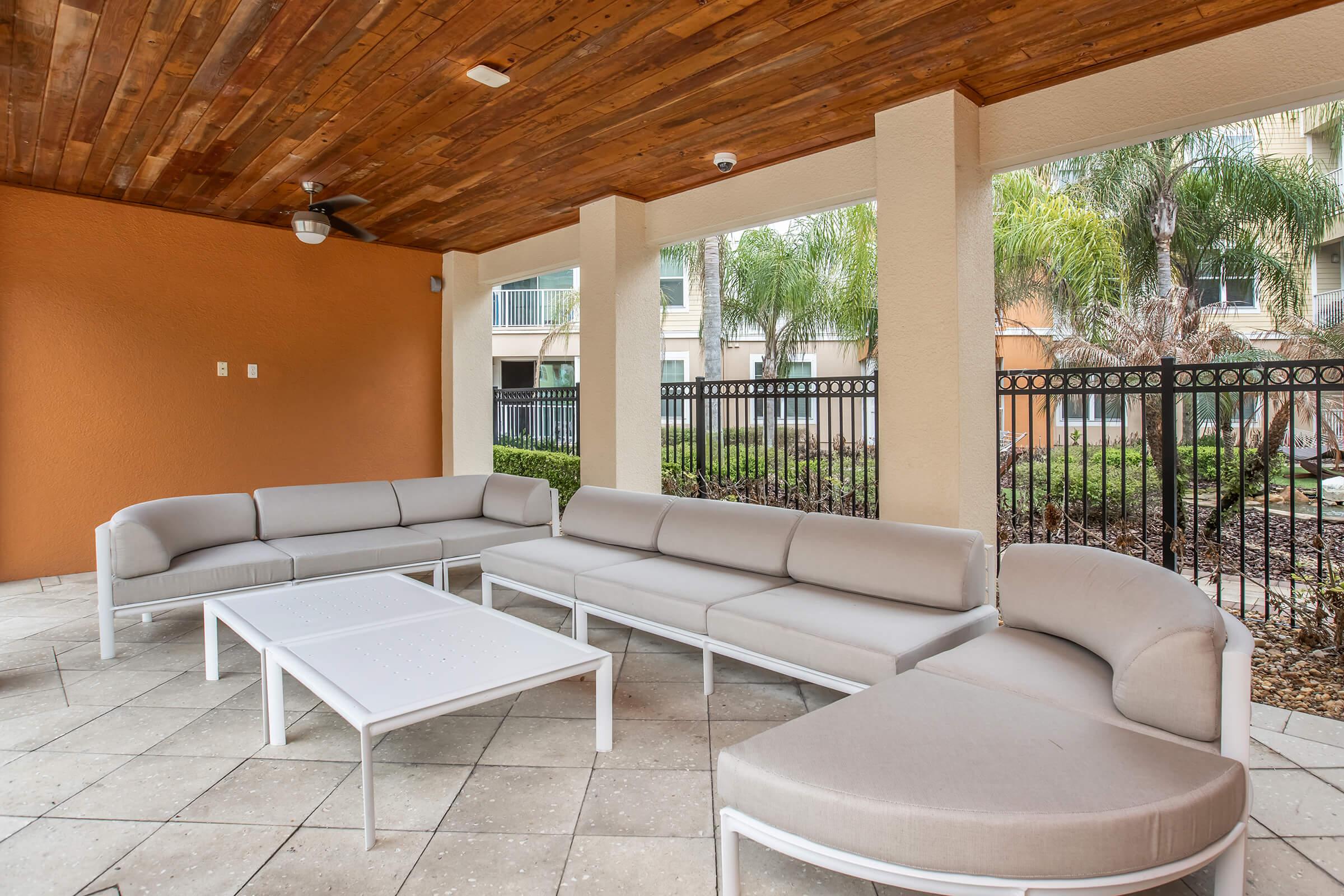 A cozy outdoor seating area under a wooden ceiling, featuring a sectional couch and a coffee table, surrounded by greenery and palm trees. The space is enclosed by a decorative fence, creating a private and inviting atmosphere.
