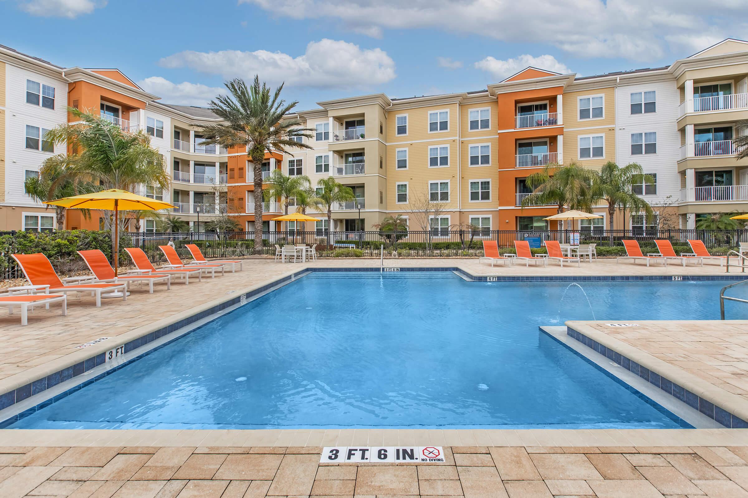 A well-maintained swimming pool surrounded by lounge chairs with orange cushions, palm trees, and vibrant apartment buildings in the background under a blue sky with clouds. The pool has a shallow area marked "3 FT 6 IN" and features umbrellas for shade.