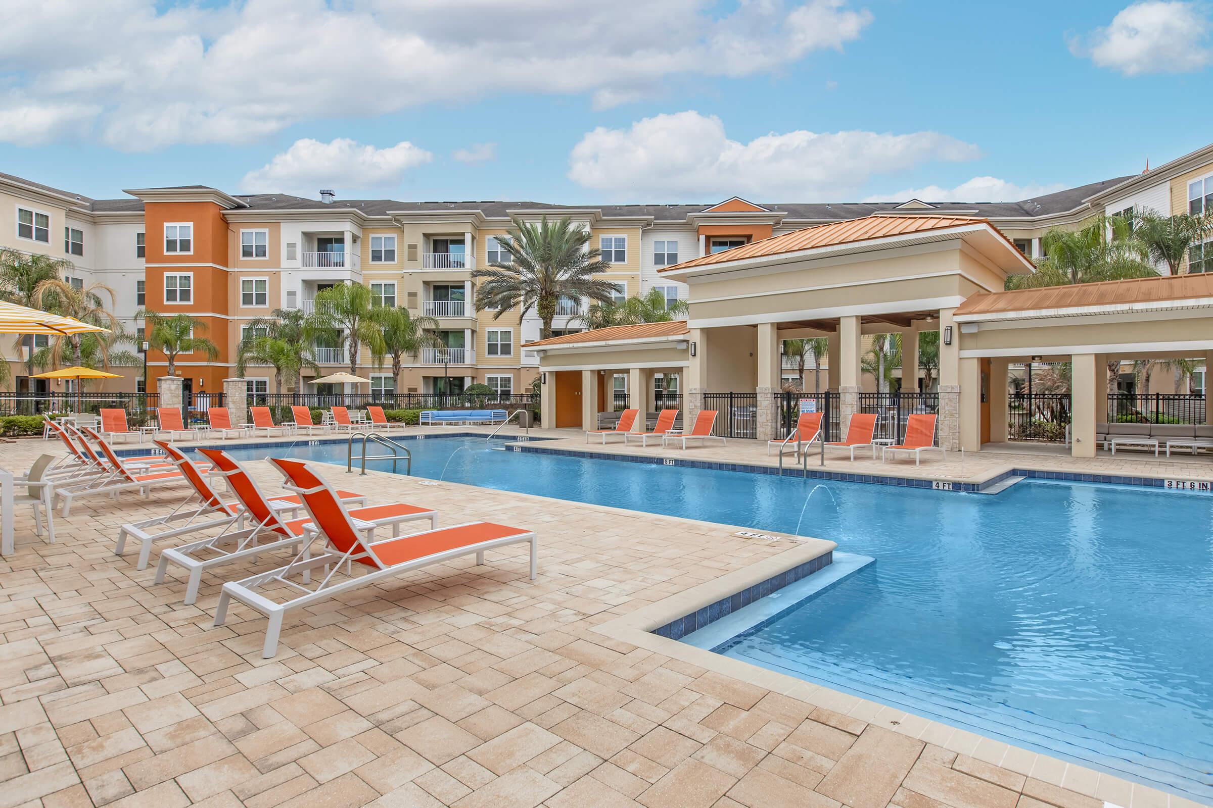 A clear blue swimming pool surrounded by orange lounge chairs and palm trees, with apartment buildings in the background. The area is sunny, featuring an awning and umbrellas for shade. The overall atmosphere is inviting and relaxing, perfect for a resort-style living experience.