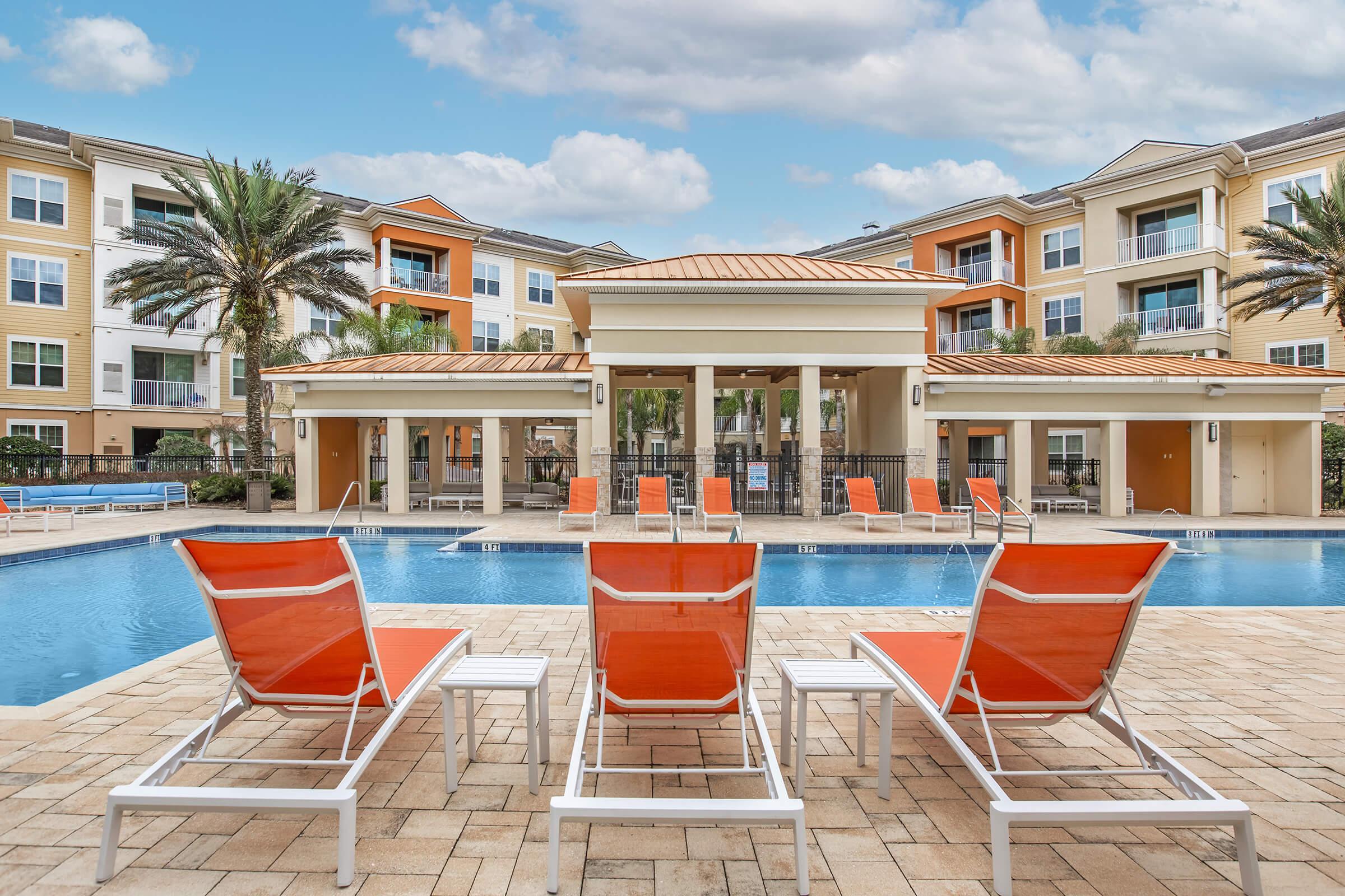 A sunny outdoor pool area featuring lounge chairs with orange cushions. In the background, there are palm trees and several apartment buildings. The clear blue water of the pool reflects the sky. The space is designed for relaxation, with a few poolside loungers arranged neatly around the pool.