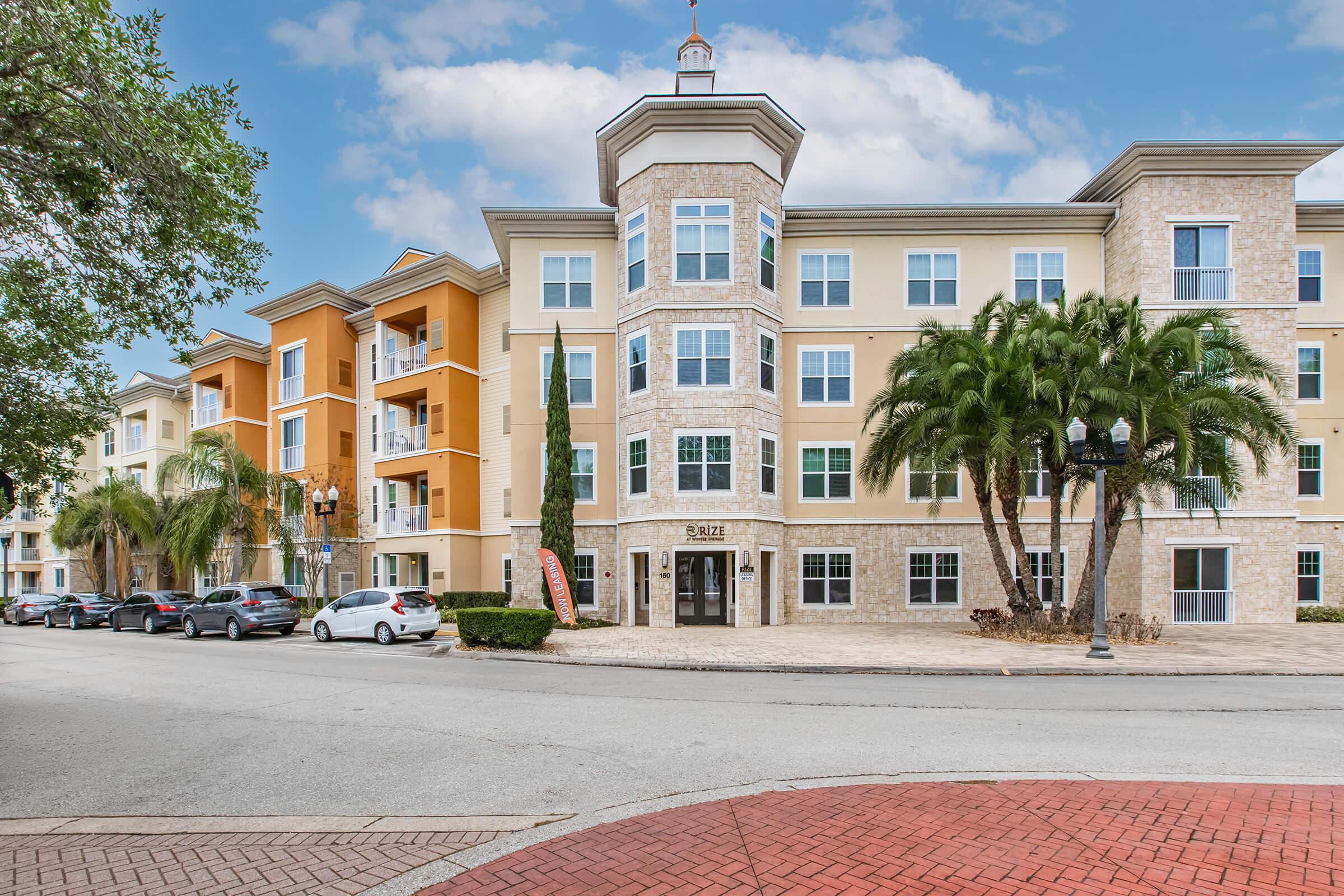 A modern apartment complex featuring a combination of stone and painted façades, with palm trees in the foreground. The building has multiple floors, large windows, and a decorative entrance. A circular brick area is visible in the foreground, along with parked cars and street lamps. The sky is partly cloudy.