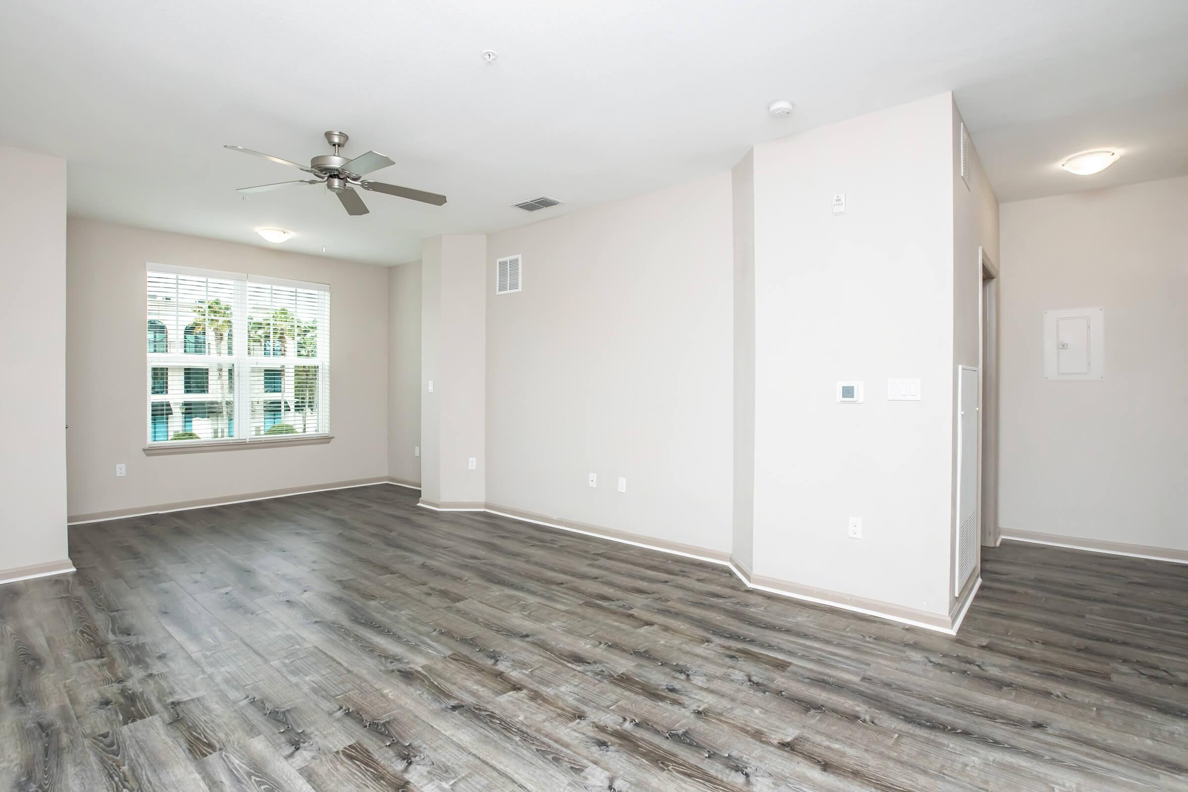 A spacious, empty living room featuring light-colored walls, wooden flooring, and a ceiling fan. Large windows allow natural light to flood the space, with blinds partially drawn. The room lacks furniture and has a doorway leading to another area.
